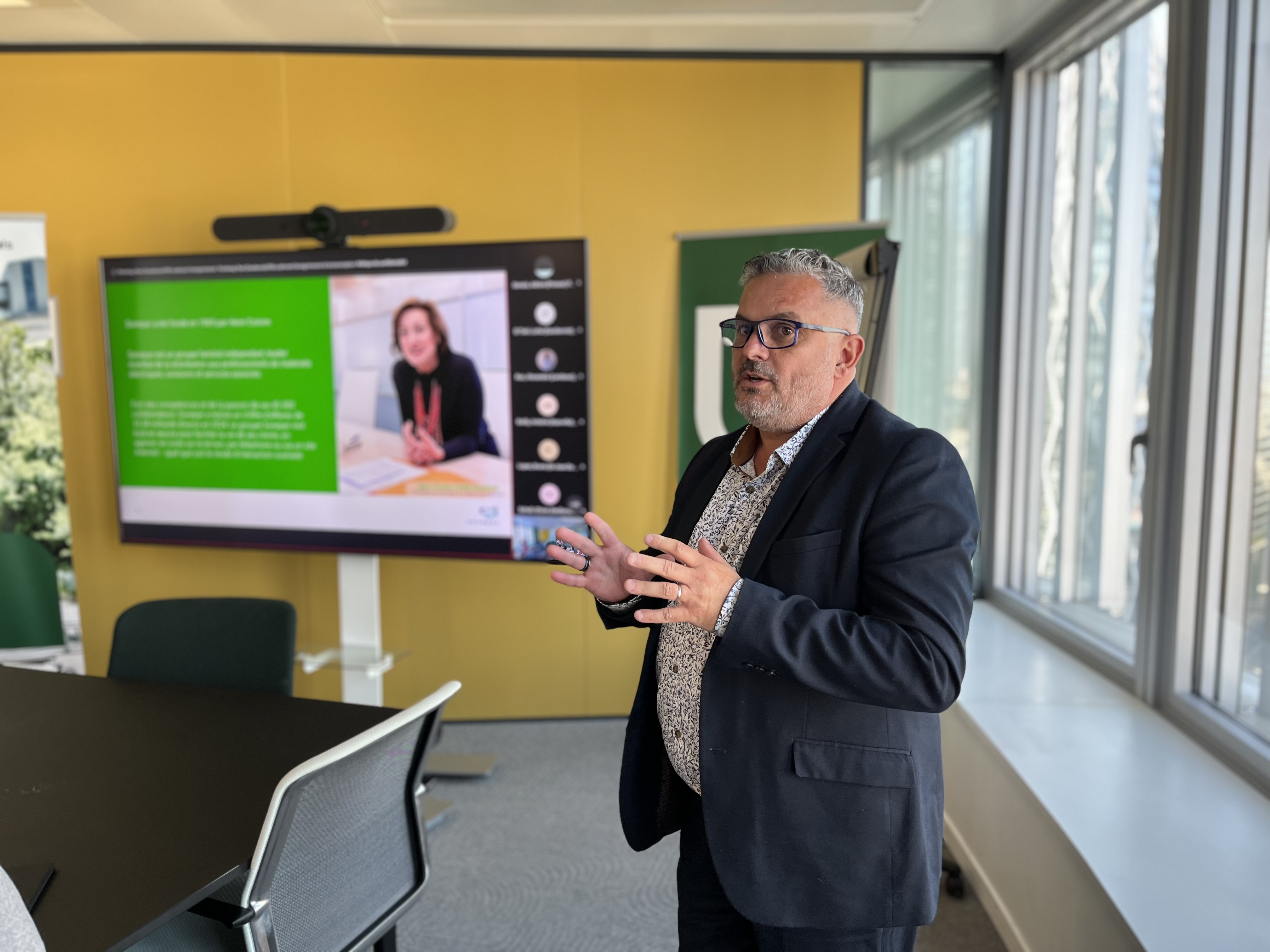 Un homme en costume parle dans une salle de réunion lumineuse avec des fenêtres. En arrière-plan, un écran affiche une présentation PowerPoint avec une diapositive contenant du texte vert, une photo d'une femme souriante et des miniatures de participants en visioconférence sur la droite. Une table et des chaises sont visibles au premier plan.
