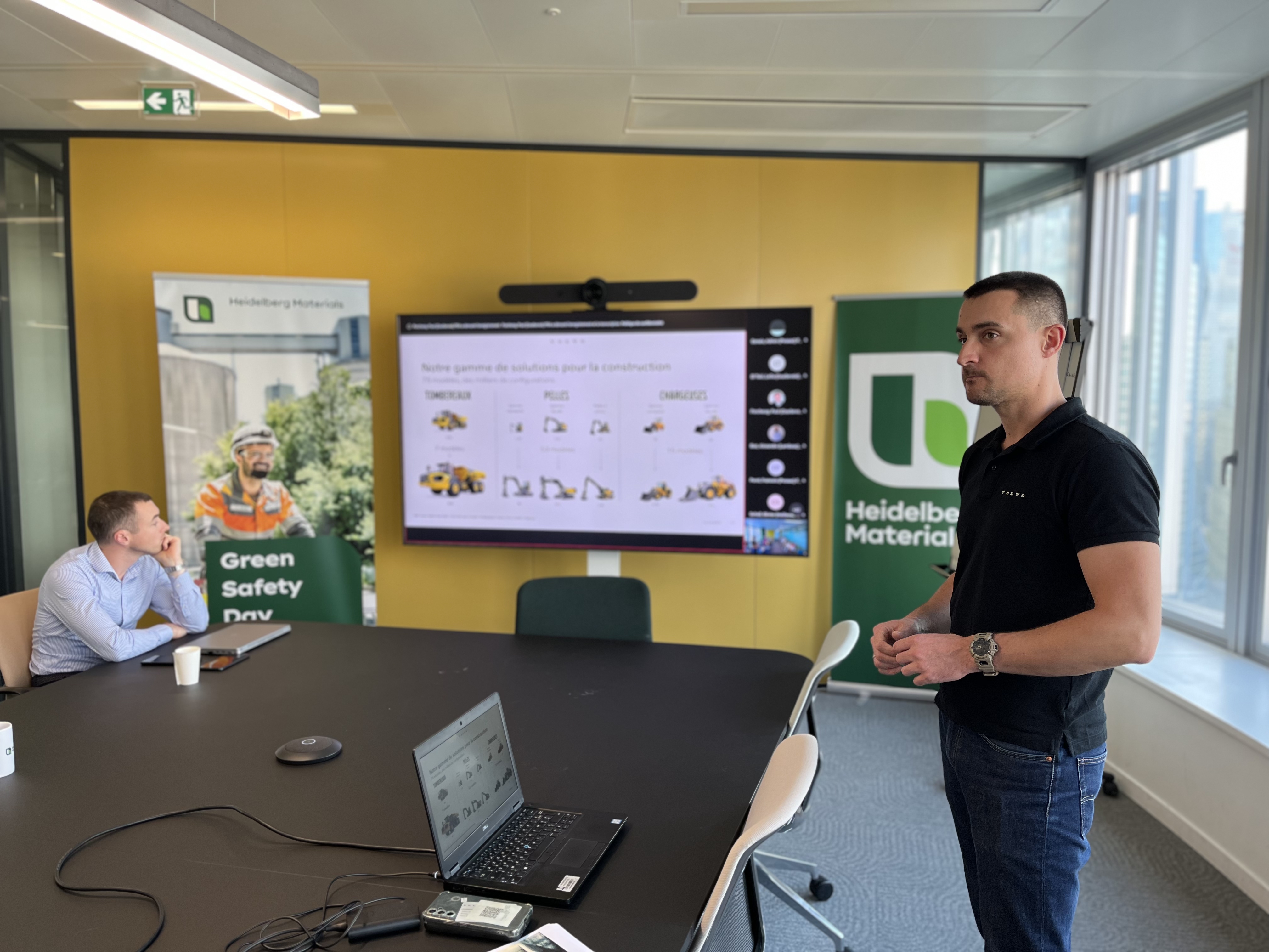 Un homme debout à droite présente un diaporama affiché sur un écran mural dans une salle de réunion lumineuse. À gauche, deux hommes sont assis devant une table noire avec des ordinateurs portables ouverts. En arrière-plan, deux affiches promotionnelles portent le logo et le nom "Heidelberg Materials", ainsi qu’une image mettant en avant la sécurité et l'environnement.