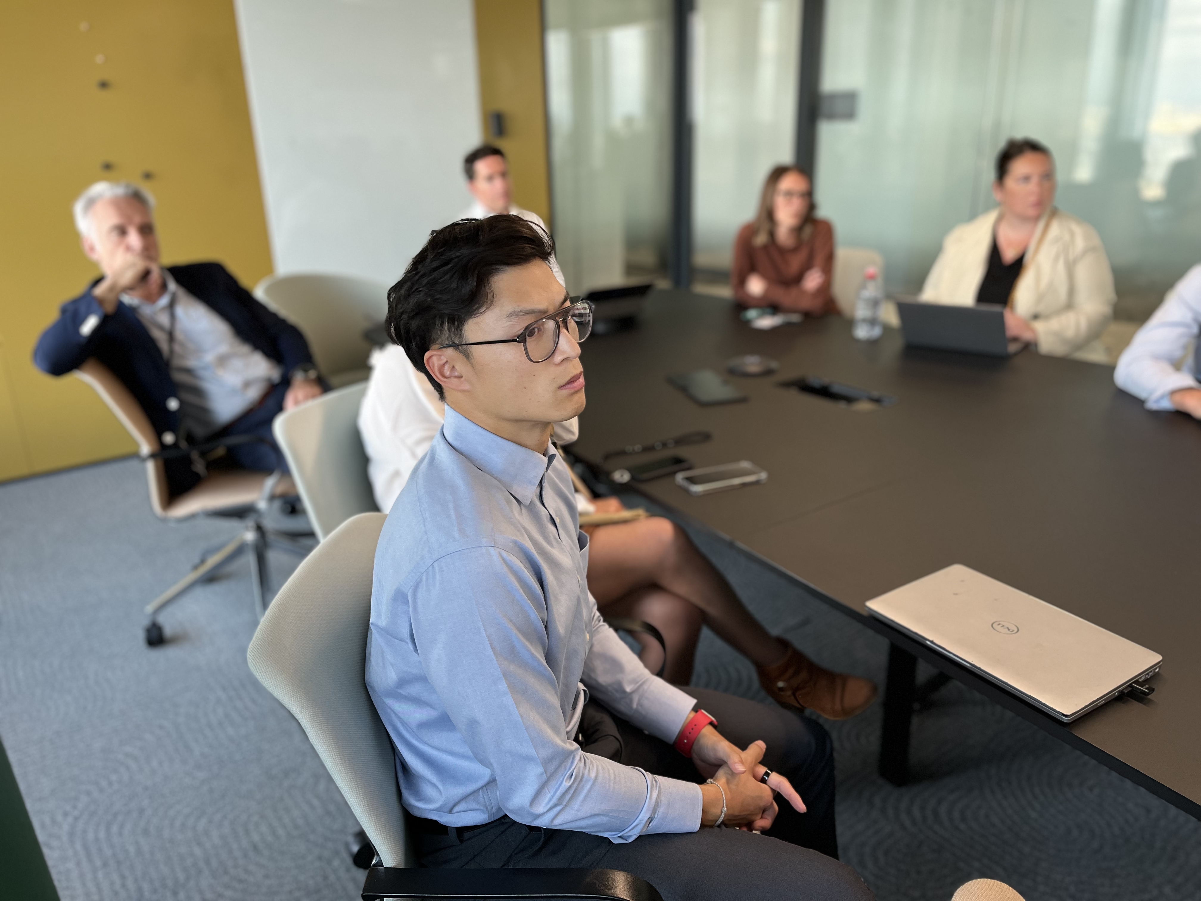 Des personnes assises autour d'une table de réunion dans un environnement de bureau moderne, certaines utilisant des ordinateurs portables. Une bouteille d'eau est visible sur la table. L'arrière-plan comprend des murs vitrés et une porte.