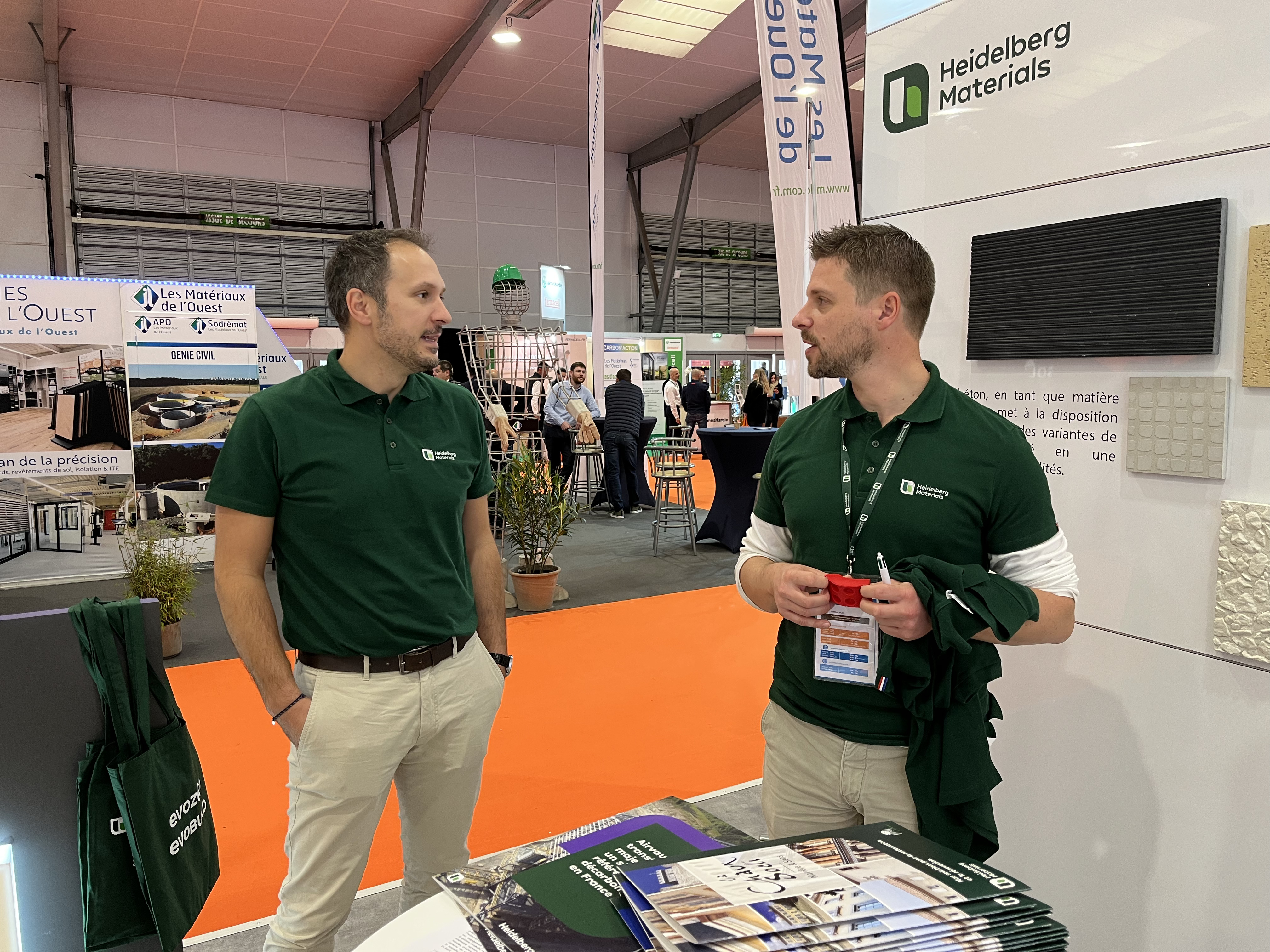 Deux hommes en polos verts avec le logo "Heidelberg Materials" discutent dans un stand d'exposition. À droite, un comptoir avec des brochures et des échantillons. En arrière-plan, d'autres stands et visiteurs dans un espace lumineux au sol orange.