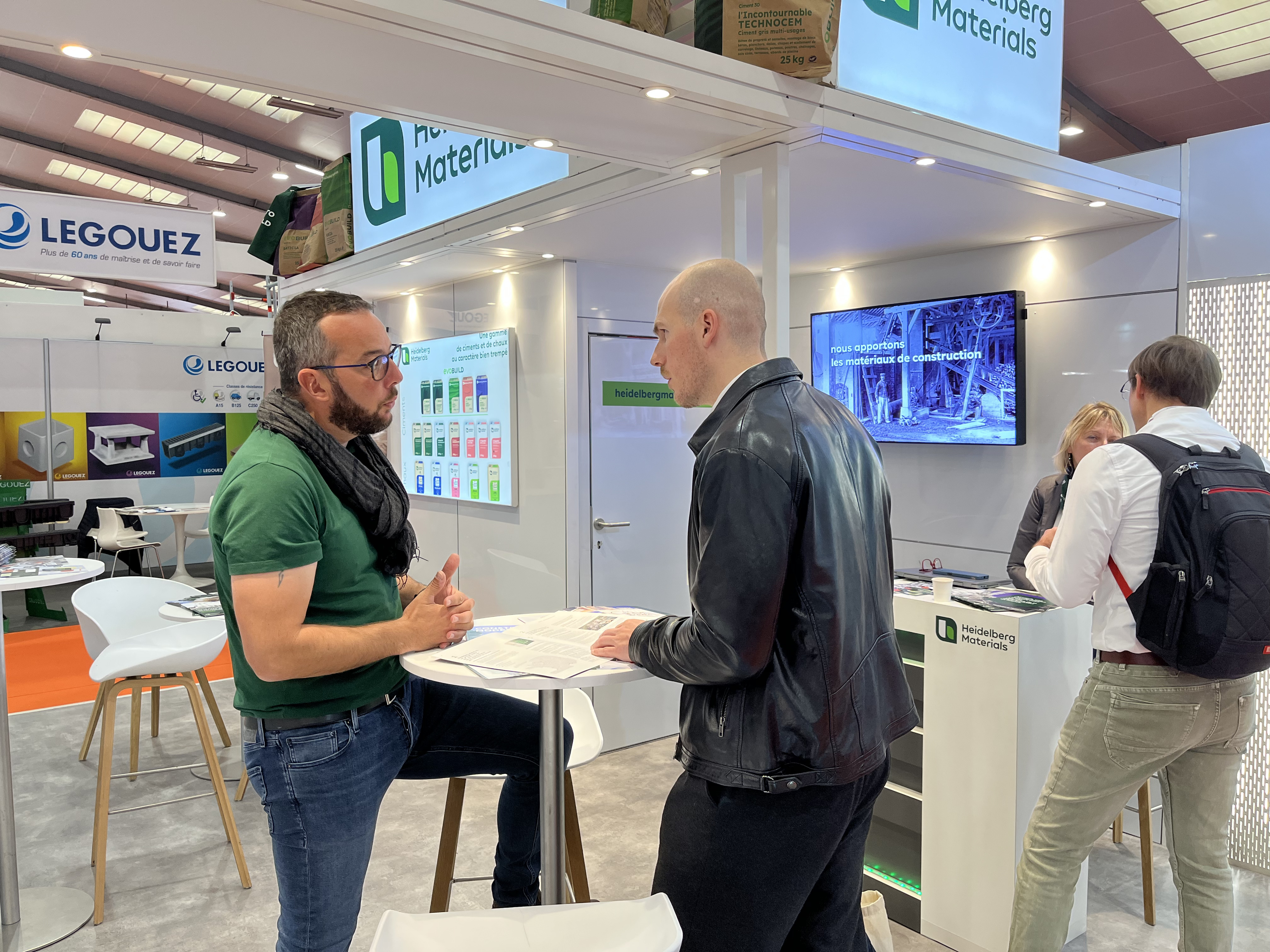 Deux hommes discutent debout devant un stand d'exposition lumineux. En arrière-plan, des panneaux affichent le logo "Molotov" et des écrans montrent des interfaces numériques. D'autres visiteurs se trouvent à droite, en train d'interagir avec les présentoirs.