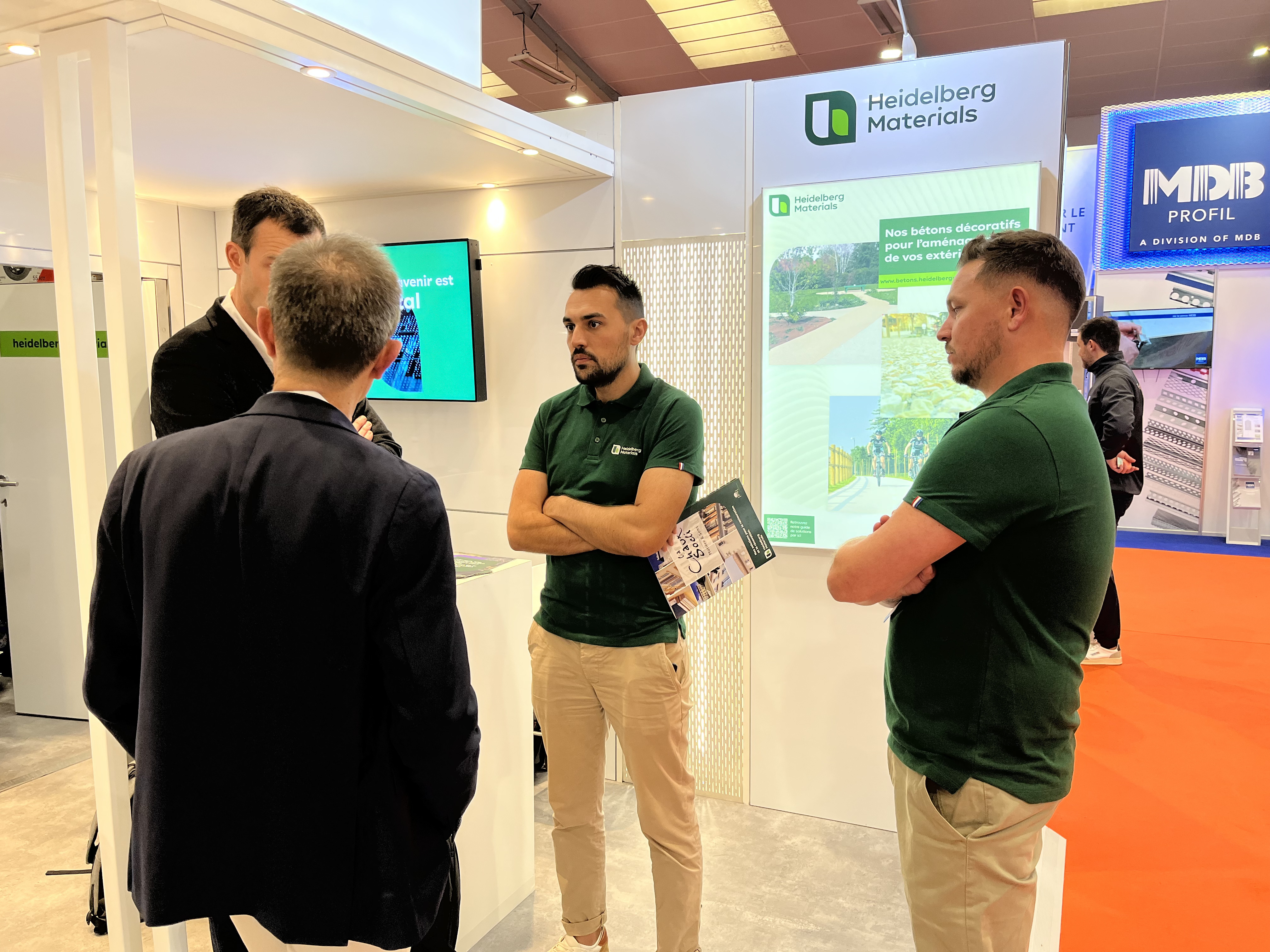 Quatre hommes discutant dans un stand d'exposition, deux portant des polos verts avec le logo "Heidelberg Materials". À l'arrière-plan, un écran et une affiche présentant les services de l'entreprise. Le sol est recouvert de moquette rouge, et d'autres stands sont visibles en arrière-plan.