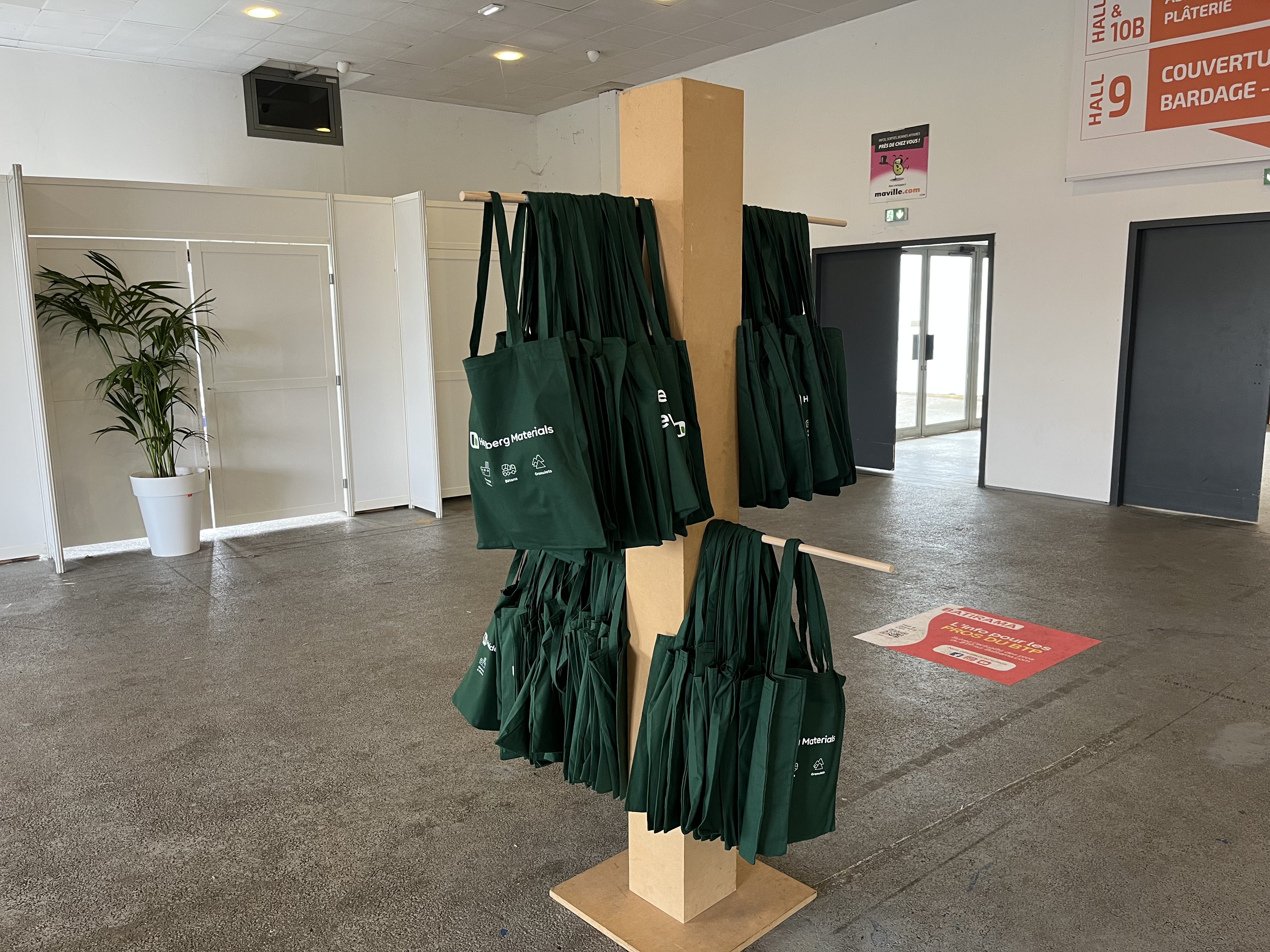 Présentoir en bois au centre d'une pièce, portant plusieurs sacs verts réutilisables accrochés sur des crochets. En arrière-plan, murs blancs avec une plante en pot à gauche, une porte vitrée au fond à droite et des panneaux d'indication rouge et blanc suspendus au plafond.