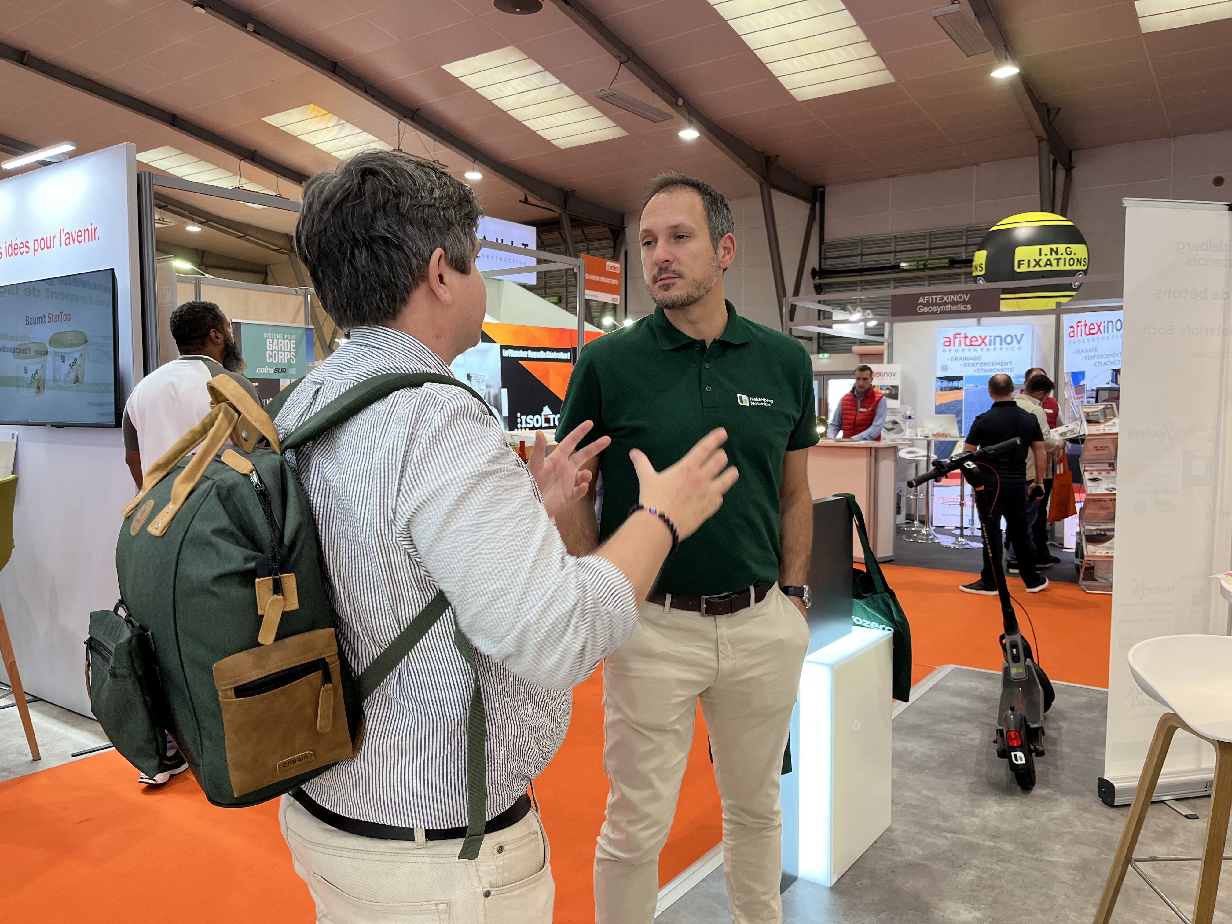 Deux hommes discutant dans un salon professionnel. L’homme à gauche porte un sac à dos vert, tandis que celui à droite, en polo vert, semble expliquer quelque chose avec des gestes. En arrière-plan, des stands d’exposition avec des affiches et des produits visibles, notamment un scooter exposé au milieu. Ambiance lumineuse et professionnelle.