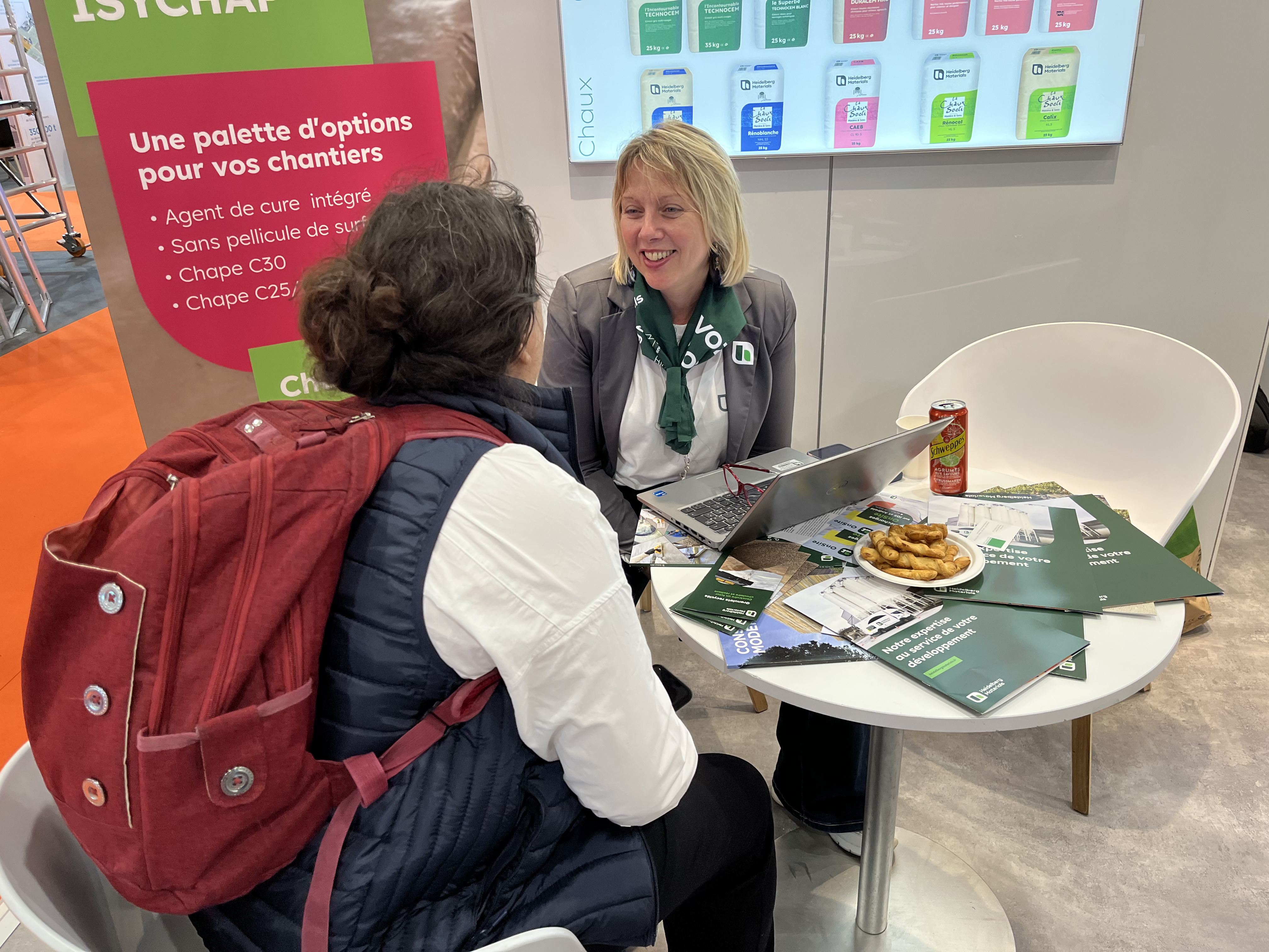 Deux femmes discutent assises à une table ronde blanche lors d'un salon professionnel. L'une porte un sac à dos rouge, tandis que l'autre, en uniforme d'entreprise avec un badge, conseille la visiteuse. Sur la table, il y a des brochures, un ordinateur portable, des biscuits et une canette. Un panneau explicatif et un écran figurent en arrière-plan, présentant des options colorées et un texte en français.