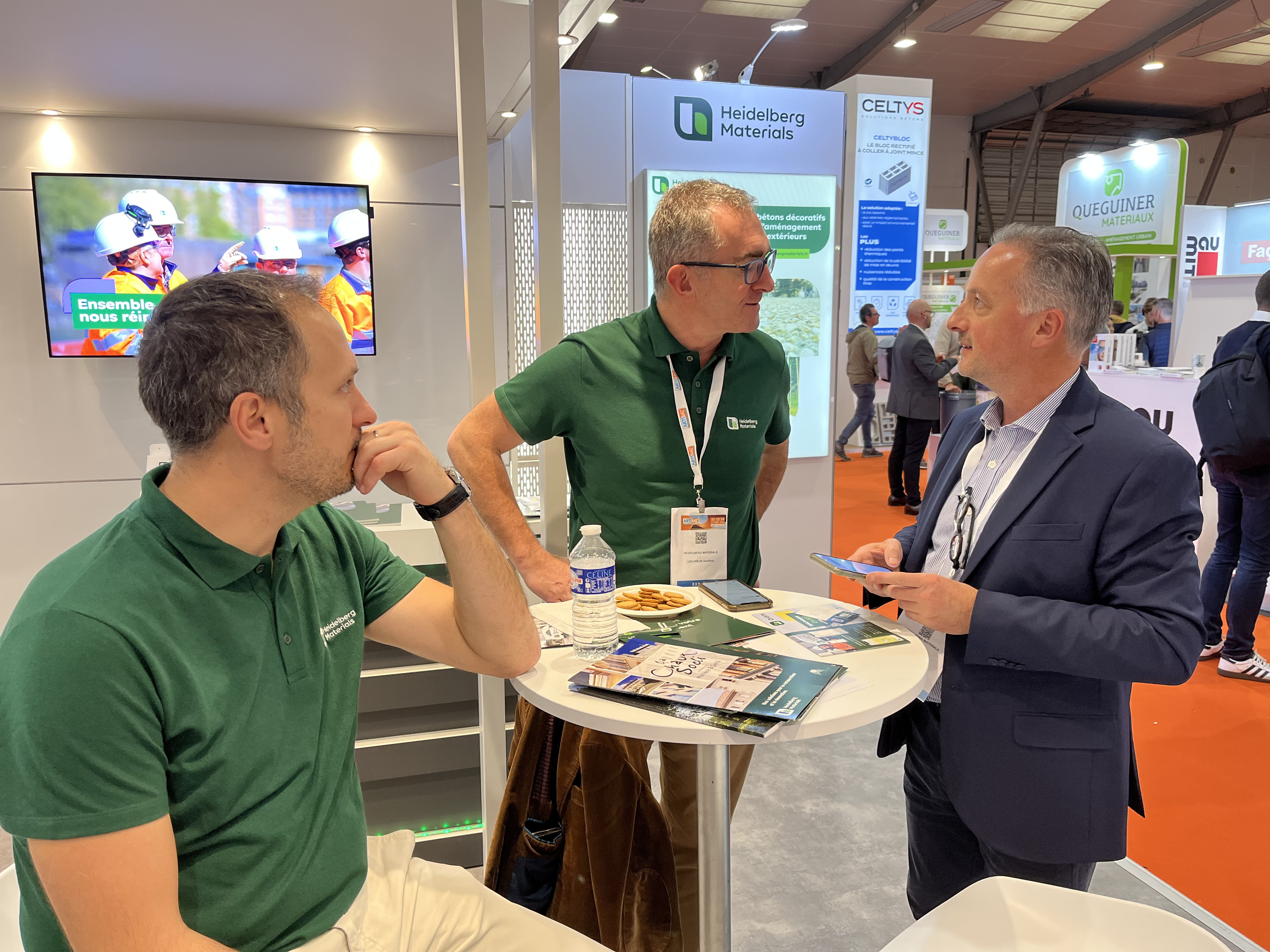 Trois hommes discutent autour d'une table haute dans un stand d'exposition. Deux d'entre eux portent des polos verts avec un logo, et l'autre est en costume-cravate. Sur la table se trouvent des brochures, un sandwich et une bouteille d'eau. En arrière-plan, un écran affiche une vidéo et des panneaux promotionnels sont visibles.