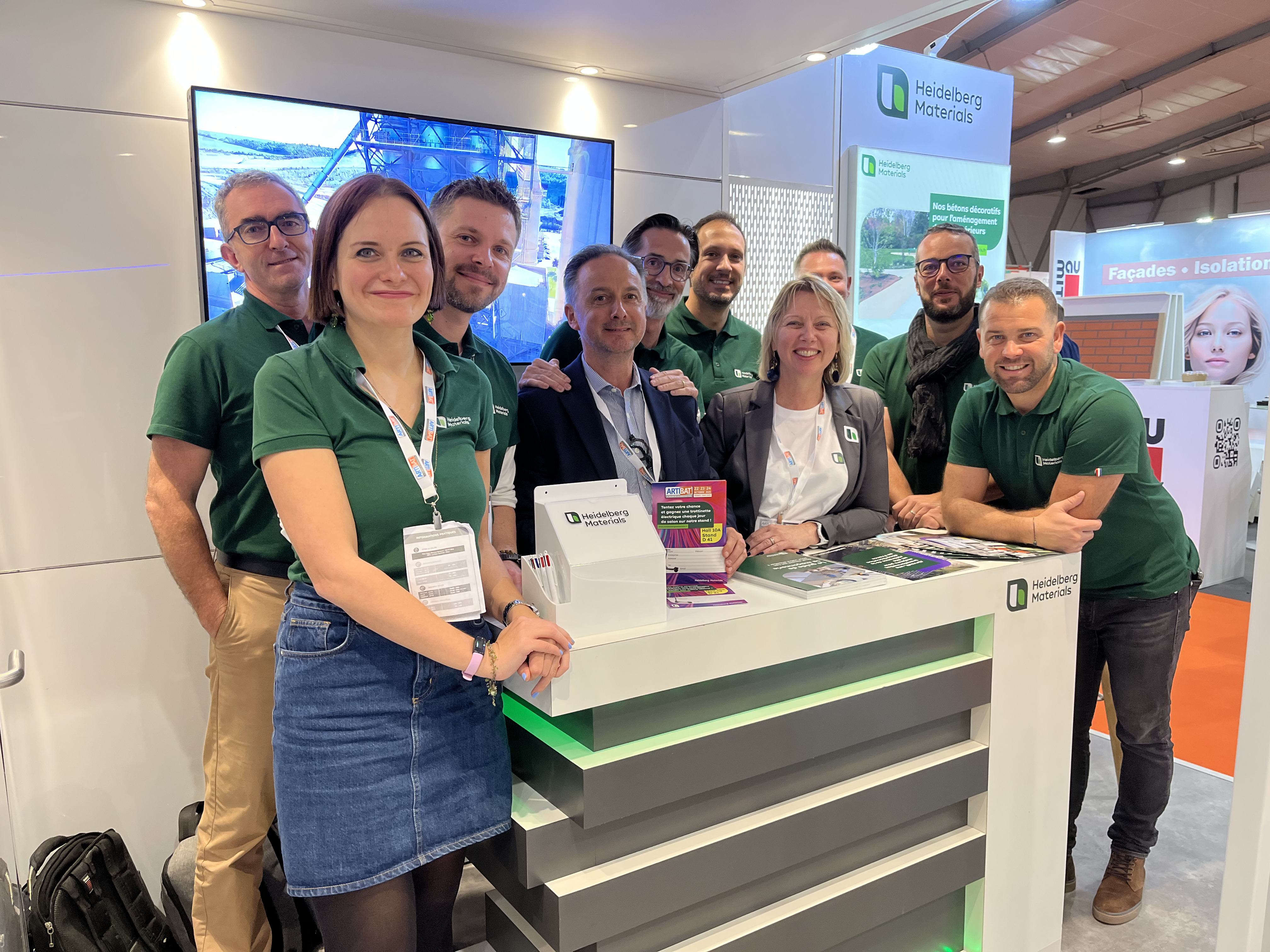 Un groupe de neuf personnes souriantes porte des polos verts autour d'un stand d'exposition moderne avec un comptoir lumineux. Sur le comptoir, des brochures, un support de cartes de visite et des objets promotionnels sont visibles. En arrière-plan, un écran affiche un bureau informatique, et des panneaux publicitaires présentent une marque. L'environnement suggère un salon professionnel ou un événement marketing.