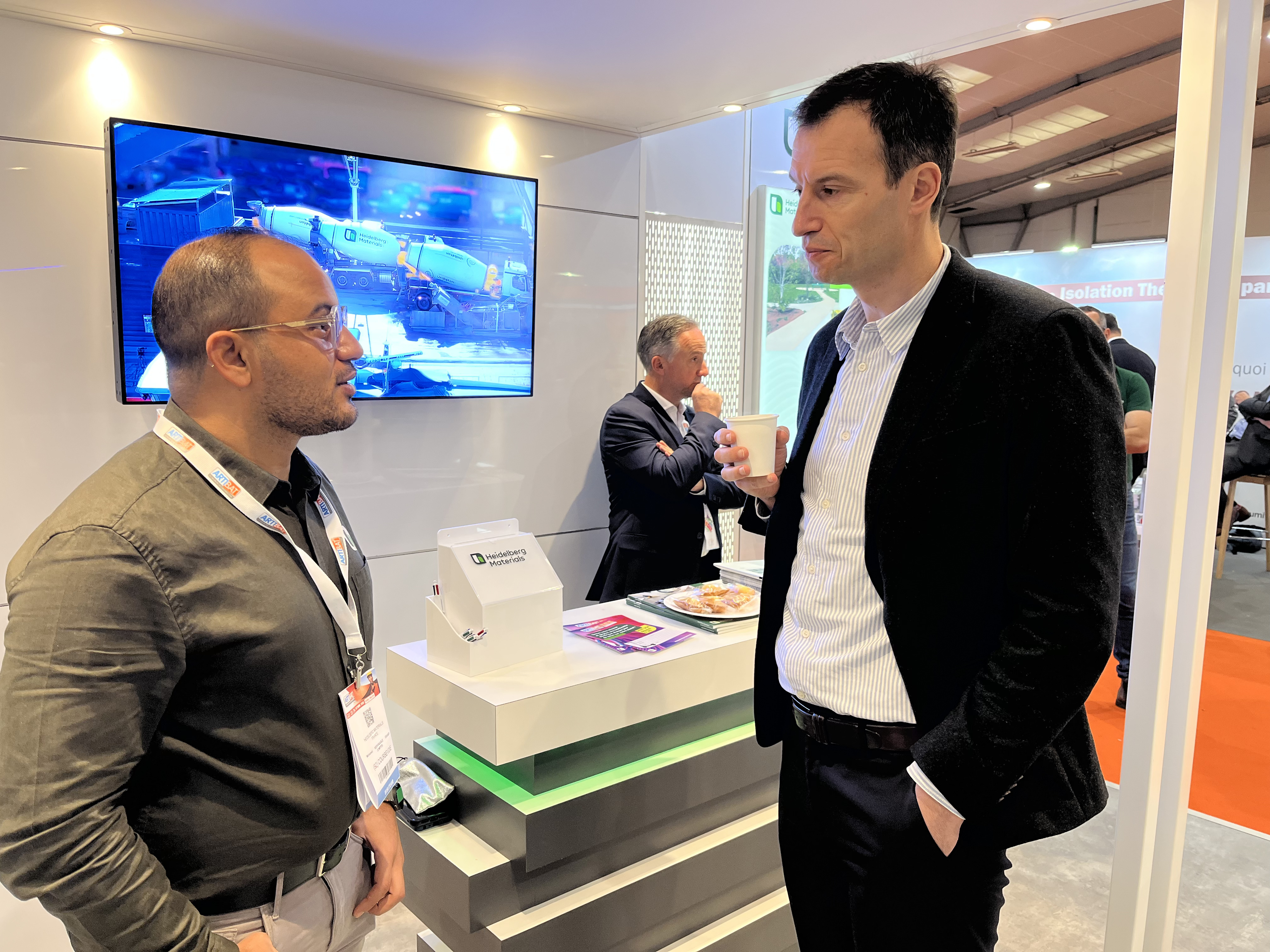 Deux hommes discutent devant un stand d'exposition moderne avec un comptoir lumineux et des brochures. En arrière-plan, un écran affiche une vidéo, et un autre homme tient une tasse tout en regardant le stand. Le sol est partiellement rouge sur la droite.