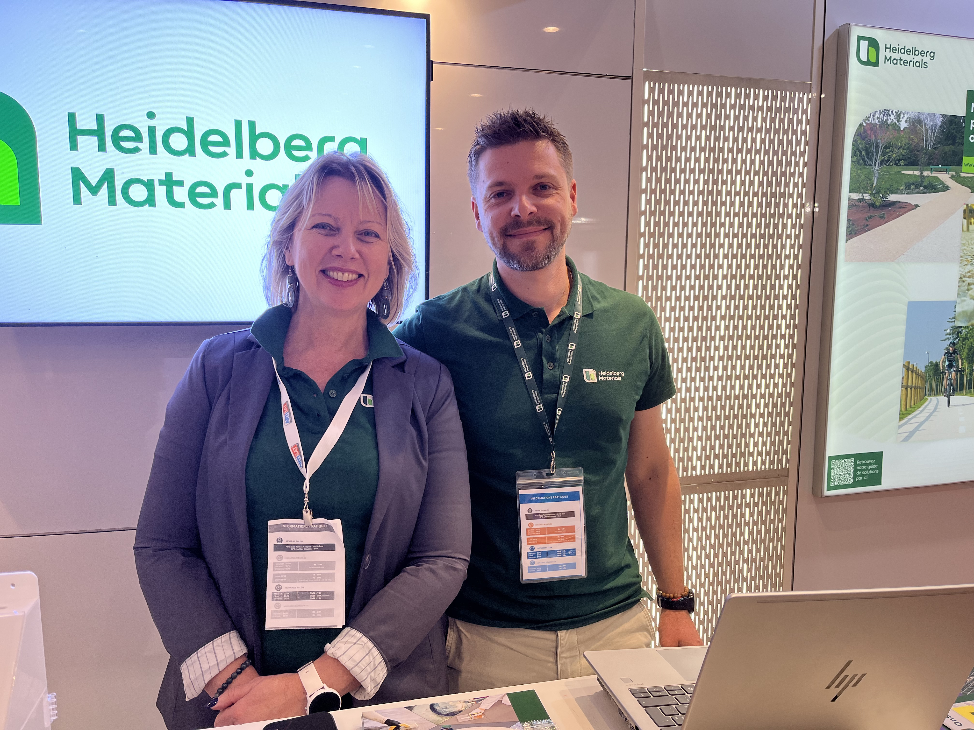 Deux personnes debout à un stand d'exposition de Heidelberg Materials. En arrière-plan, un écran affiche le logo de l'entreprise sur la gauche et une image promotionnelle d'un paysage urbain sur la droite. Une table au premier plan contient un ordinateur portable et des documents.