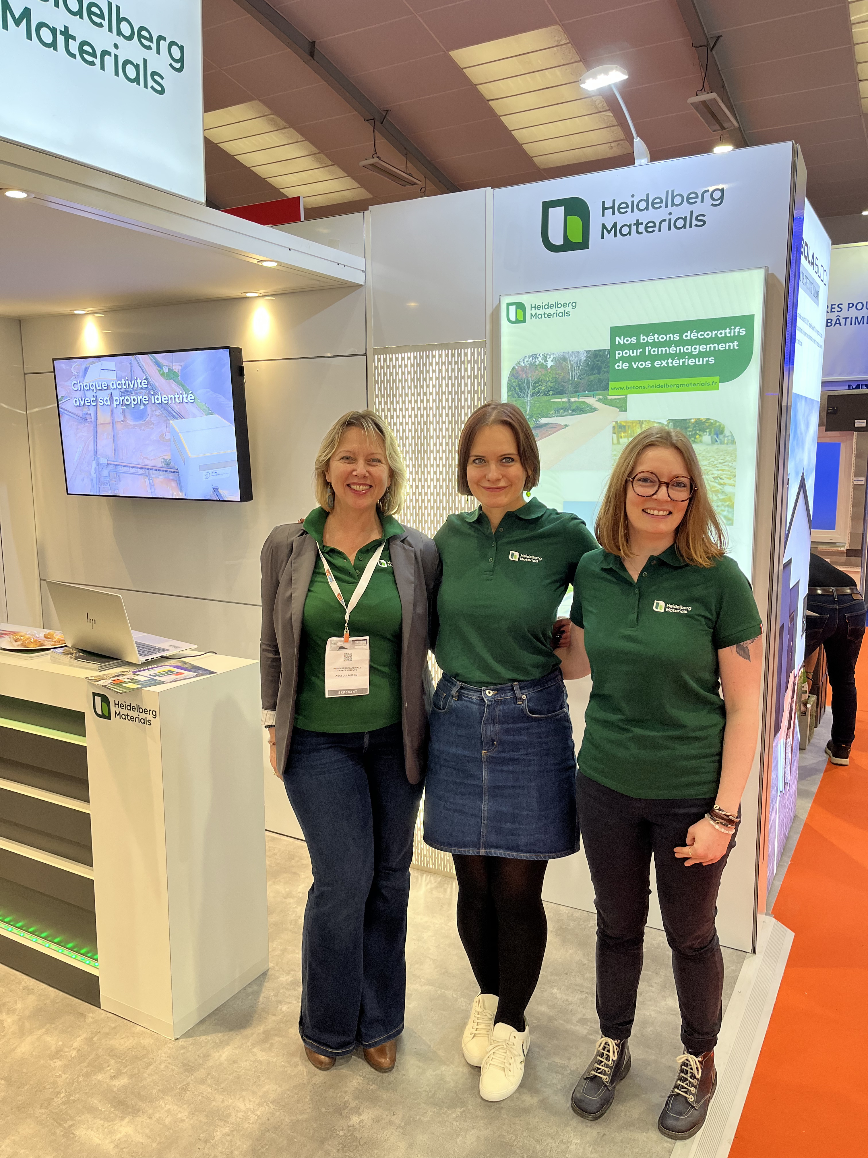 Trois femmes souriantes debout devant un stand d'exposition de Heidelberg Materials. Elles portent des polos verts avec le logo de l'entreprise. En arrière-plan, un écran affiche une carte interactive et un panneau présente des informations sur les matériaux et solutions durables. Le sol est gris, et le stand a une apparence moderne aux teintes blanches.