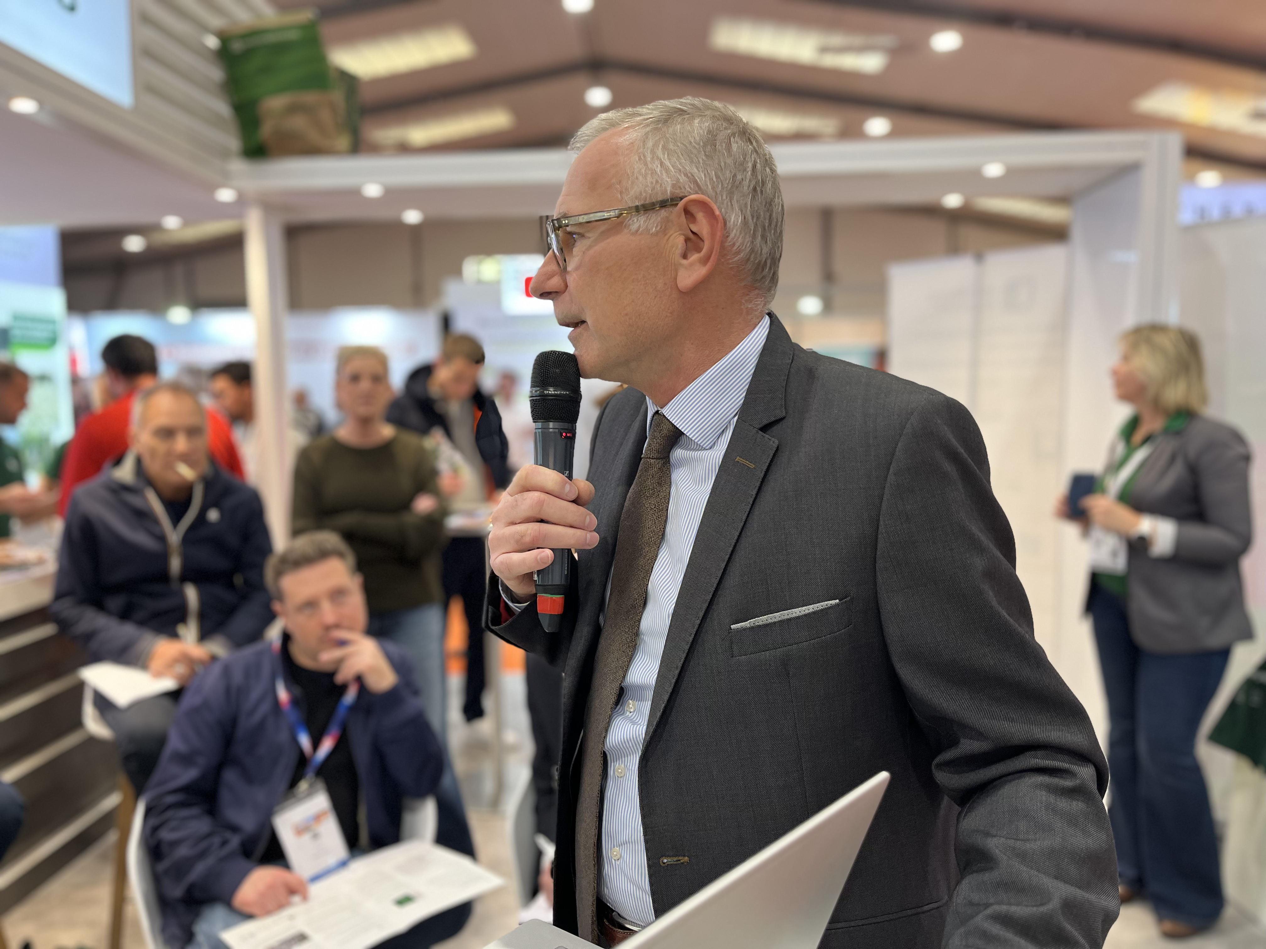 Un homme en costume tenant un microphone donne une présentation dans un espace d'exposition, entouré de participants assis et debout. L'arrière-plan montre des stands, des affiches et des visiteurs dans un environnement professionnel.