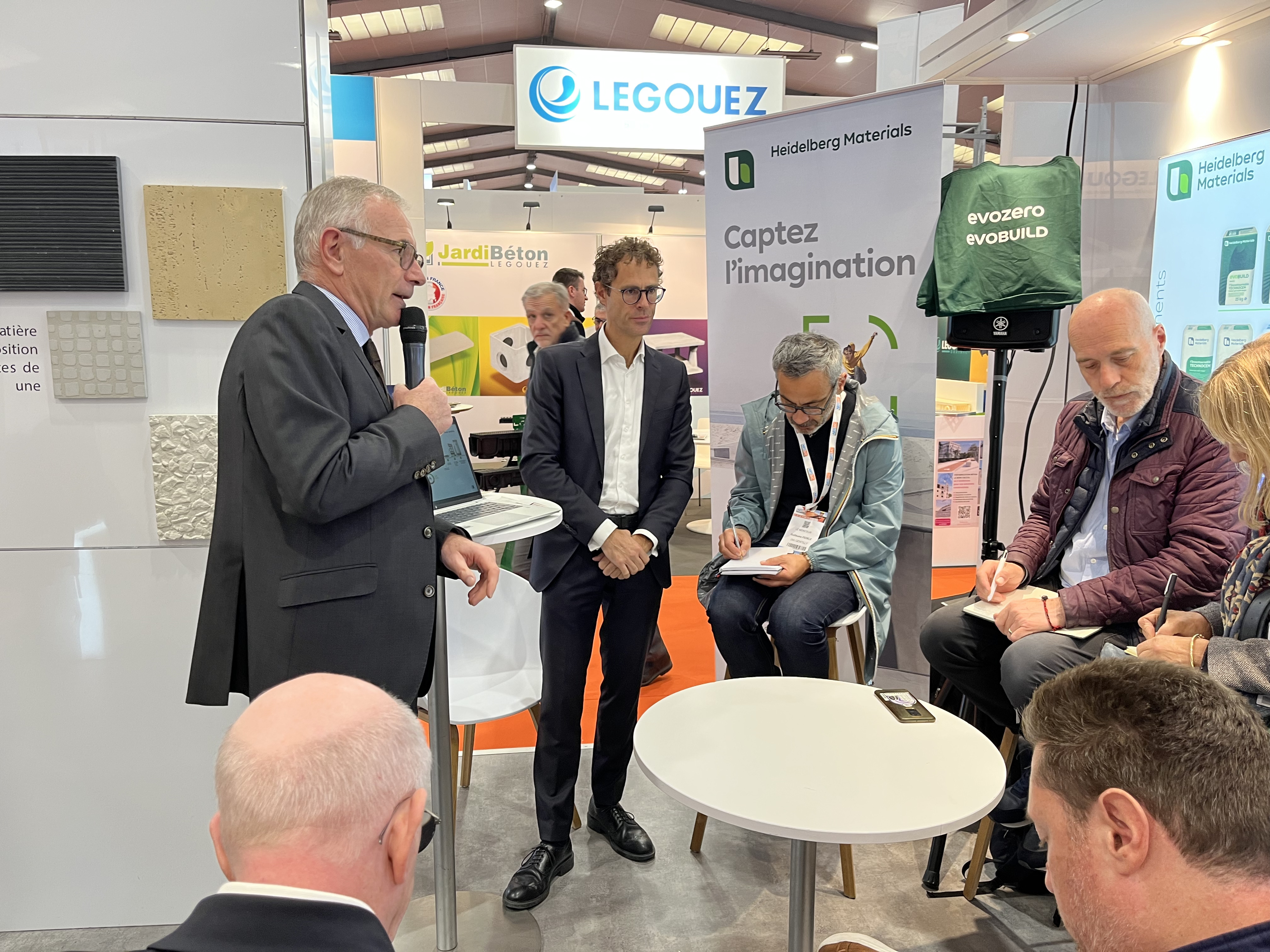 Un homme en costume parle dans un micro lors d'une présentation dans un salon professionnel, entouré de quatre personnes assises prenant des notes, près d'une table blanche. En arrière-plan, des panneaux d'affichage indiquent "Captez l'innovation" et le logo "LEGOUEZ" est visible.