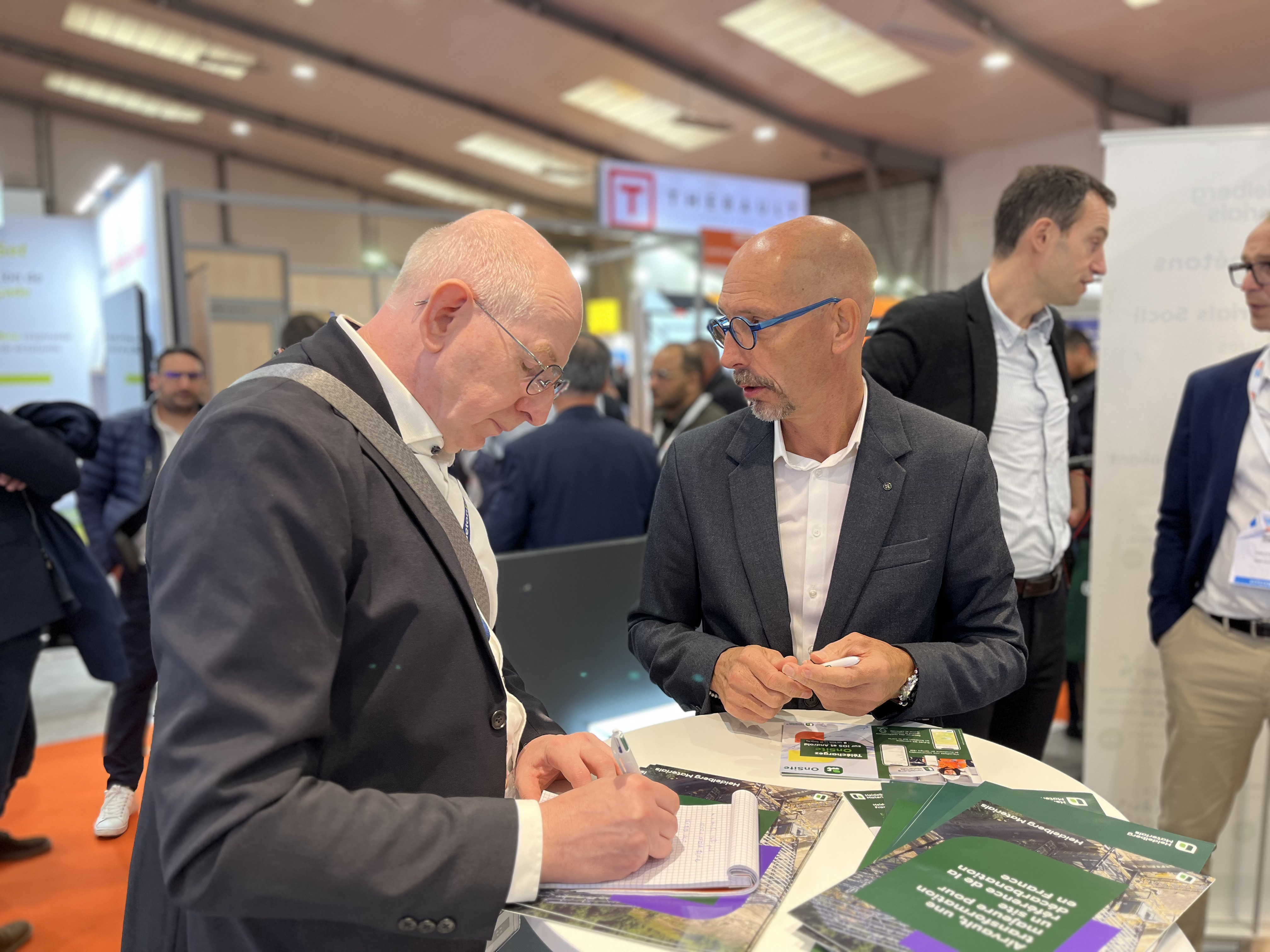 Deux hommes en costume discutent autour d’une table lors d’un salon professionnel. L’un écrit sur un carnet tandis que l’autre tient une carte. Sur la table, des brochures et des documents colorés sont visibles. À l'arrière-plan flouté, d'autres participants et des stands d'exposition sont présents.