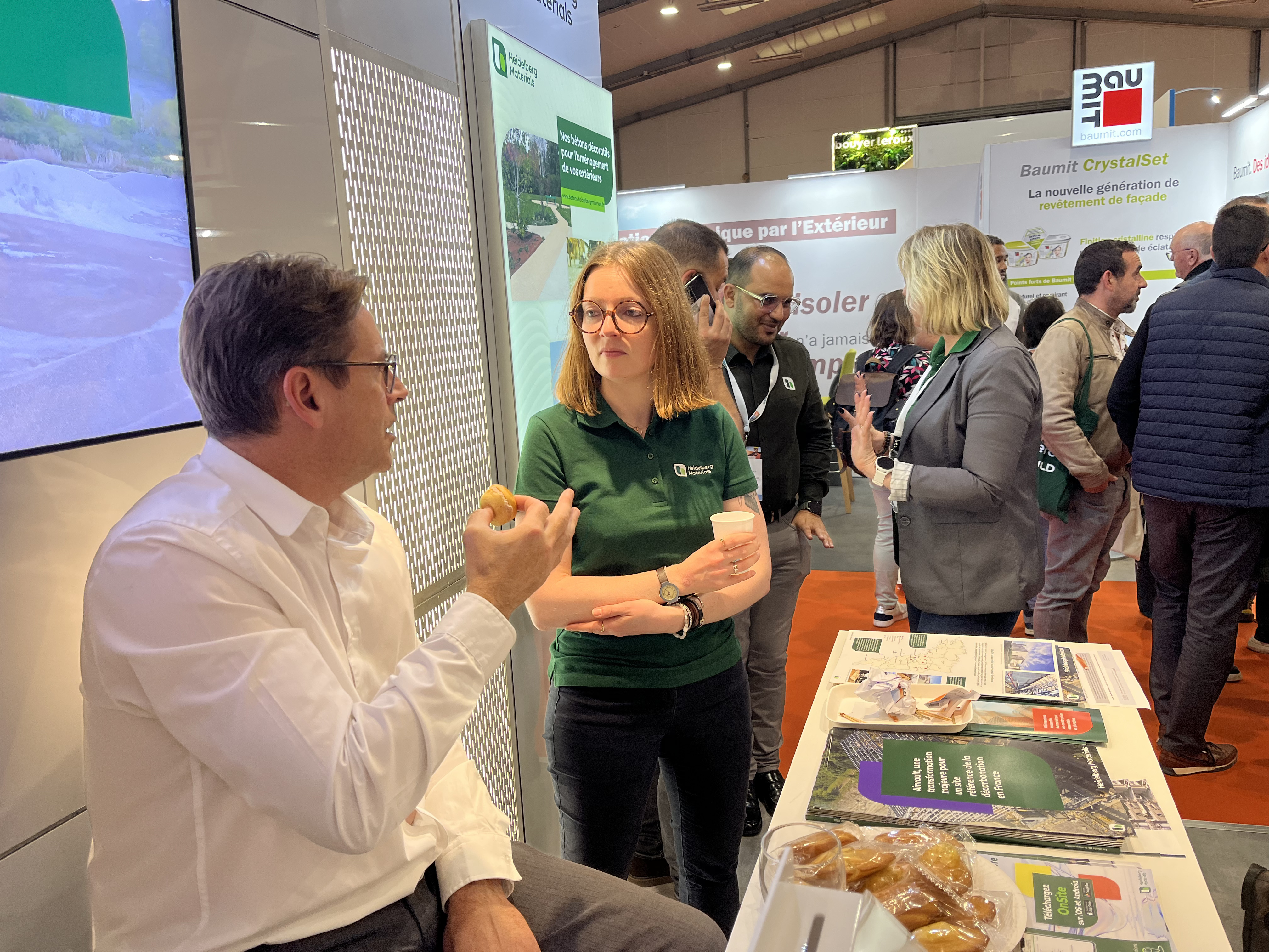 Un homme en chemise blanche discute avec une femme portant un polo vert à un stand d'exposition. Sur la table devant eux, des brochures, un téléphone avec une application visible et des viennoiseries. En arrière-plan, d'autres participants interagissent dans un espace d'exposition lumineux.