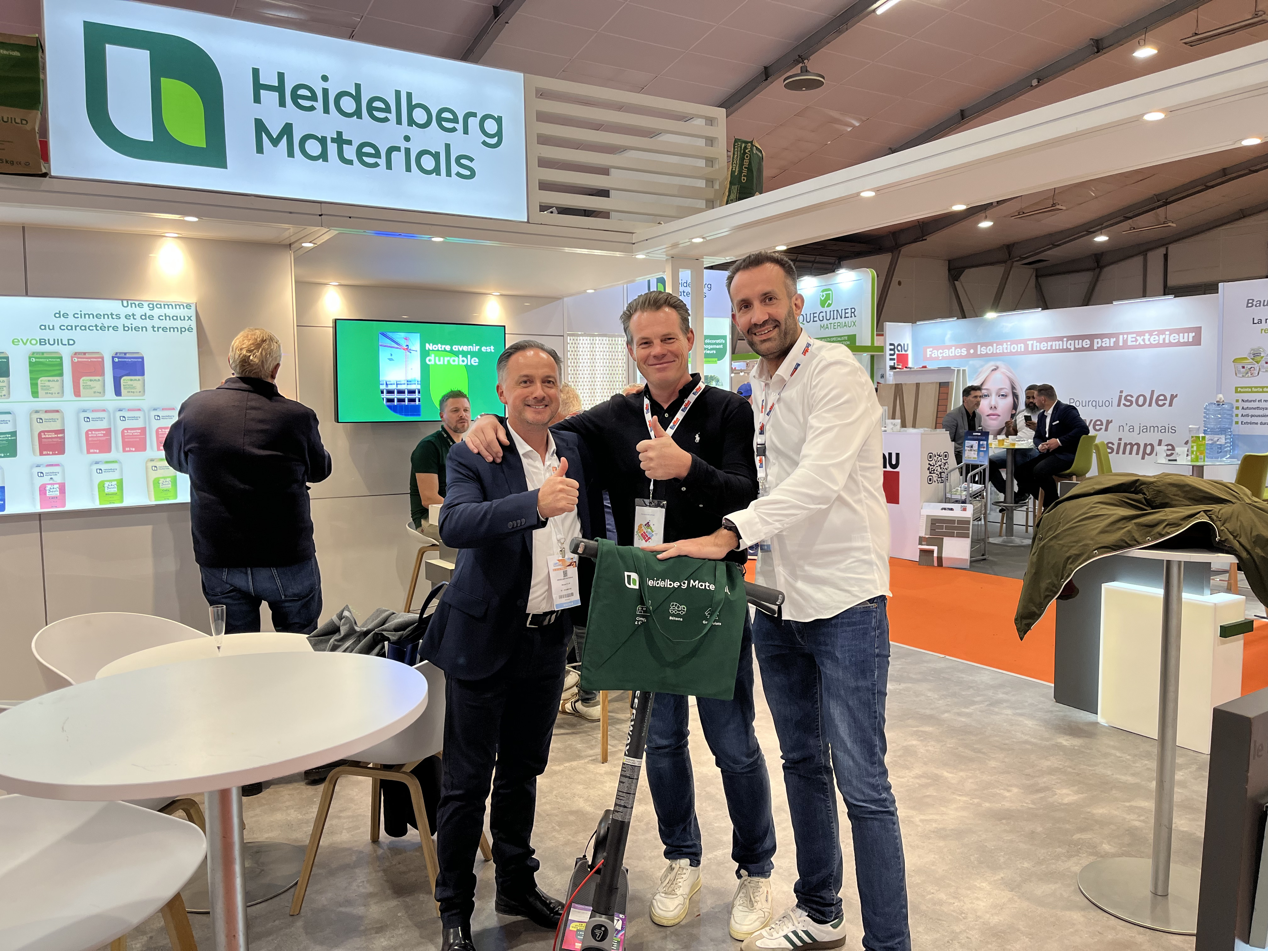 Trois hommes souriants posent devant un stand d’exposition marqué "Heidelberg Materials". L’homme au centre tient un sac vert portant le logo de l’entreprise. À l’arrière, des affiches colorées, du mobilier moderne et des visiteurs sont visibles, avec une ambiance de salon professionnel.