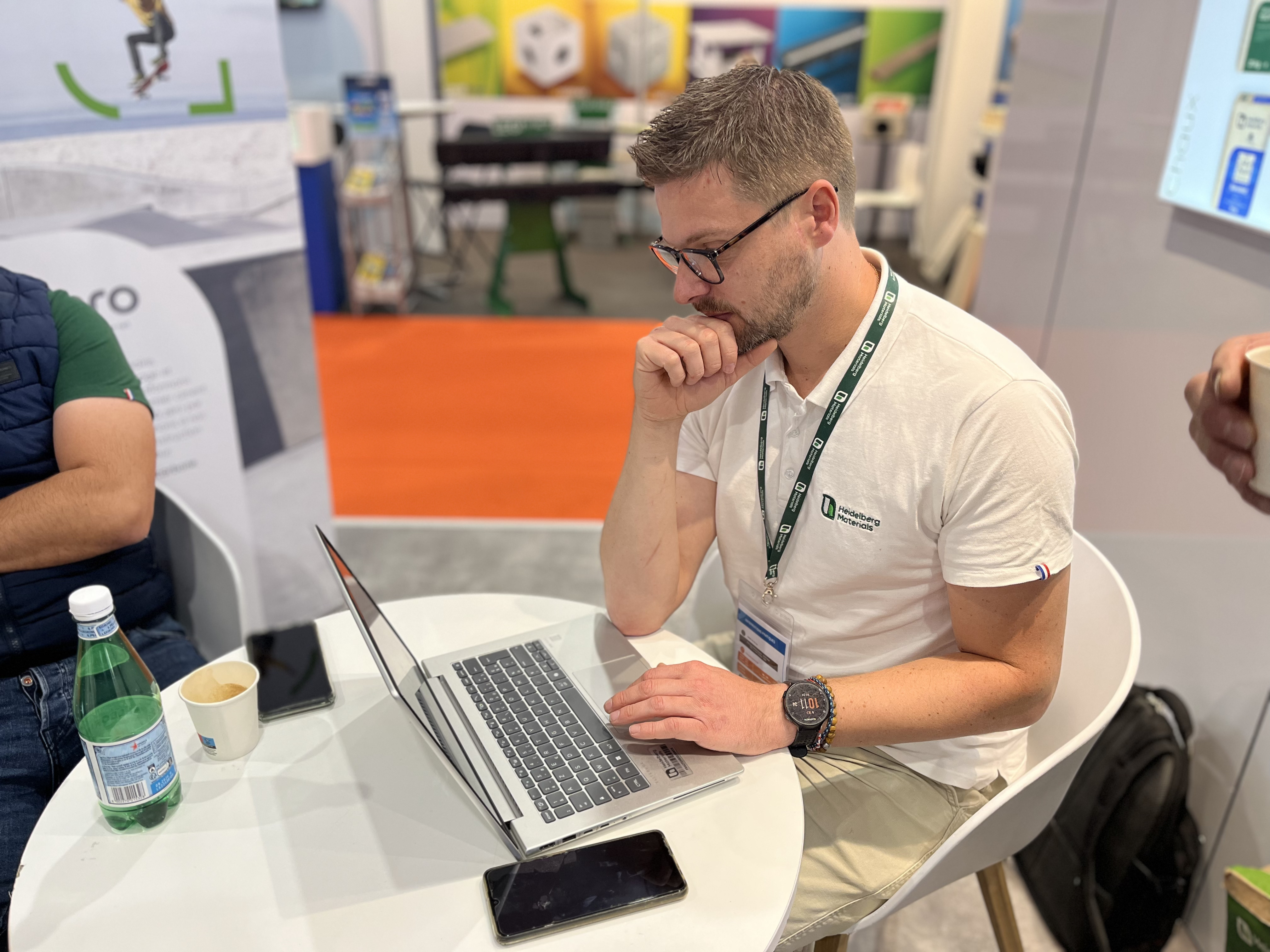Un homme assis à une table ronde utilisant un ordinateur portable, entouré d'objets comme une bouteille d'eau, une tasse et un smartphone. En arrière-plan, des panneaux colorés et des stands de présentation sont visibles dans un environnement professionnel ou d'exposition.