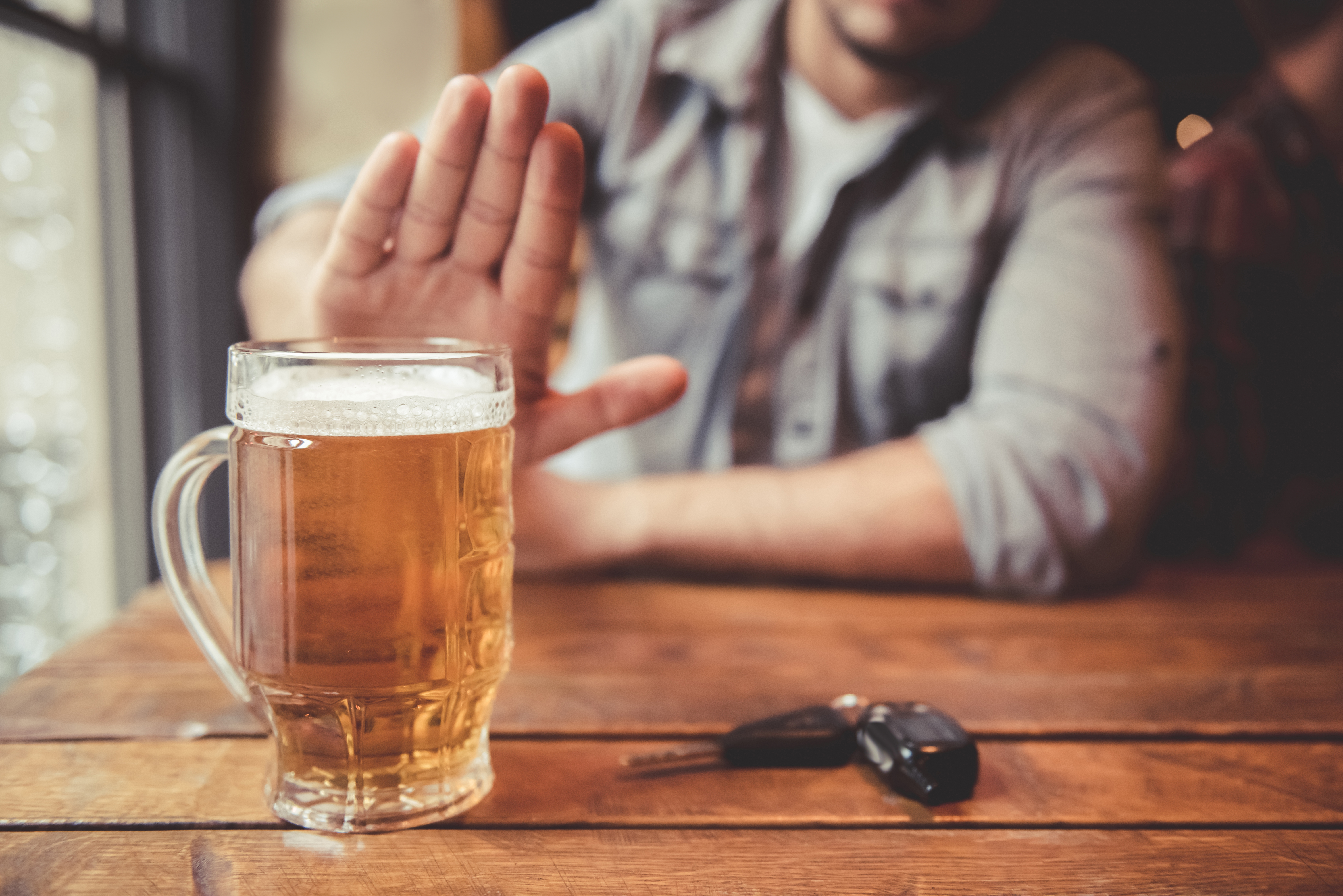 Une personne vêtue d'une chemise à carreaux tend la main dans un geste d'interdiction vers une chope de bière posée sur une table en bois ; des clés de voiture sont placées à proximité, soulignant le refus de boire et de conduire.