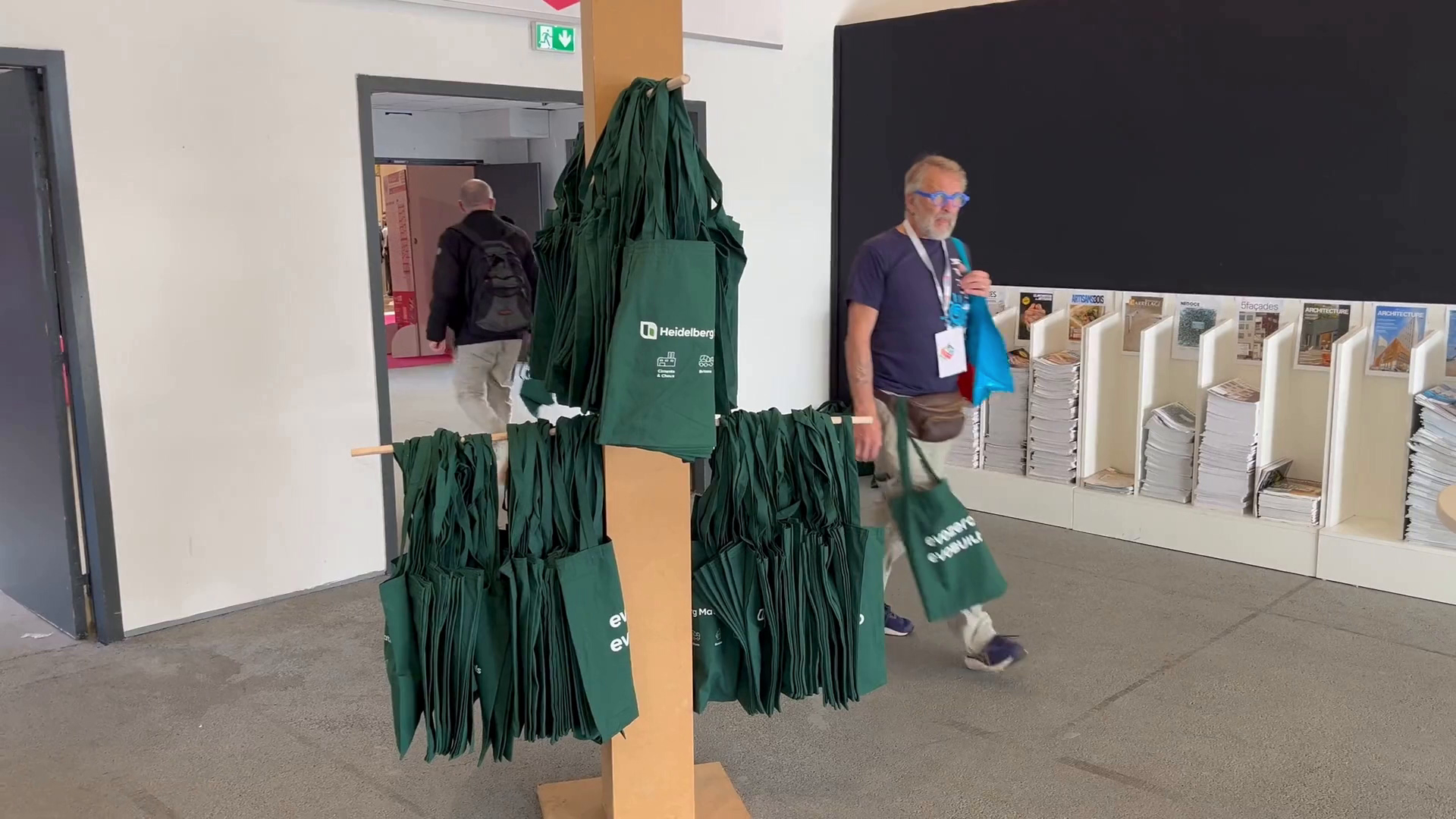 Un support en bois tient plusieurs sacs fourre-tout verts avec du texte imprimé en blanc, placés au centre. Un homme portant des lunettes, une chemise bleue et un masque facial marche vers la droite, tenant un sac fourre-tout vert et un sac bleu. À l'arrière-plan, une porte ouverte montre une autre personne qui sort, et des étagères avec des articles blancs bordent le mur de droite.
