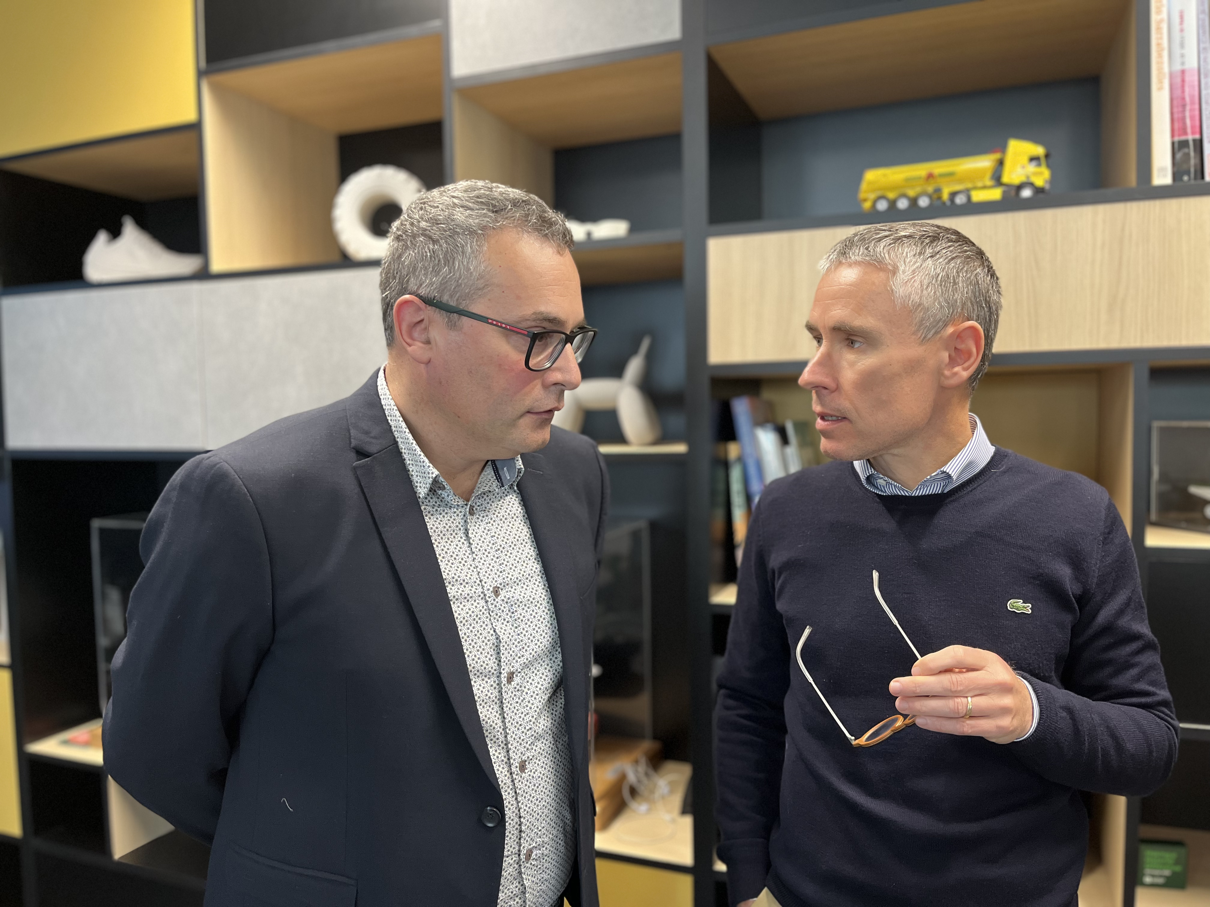 Deux hommes en tenue professionnelle discutent dans un bureau moderne, avec des étagères en arrière-plan contenant divers objets décoratifs, des livres et une miniature de camion jaune visible en haut à droite. L'un tient des lunettes dans sa main droite.
