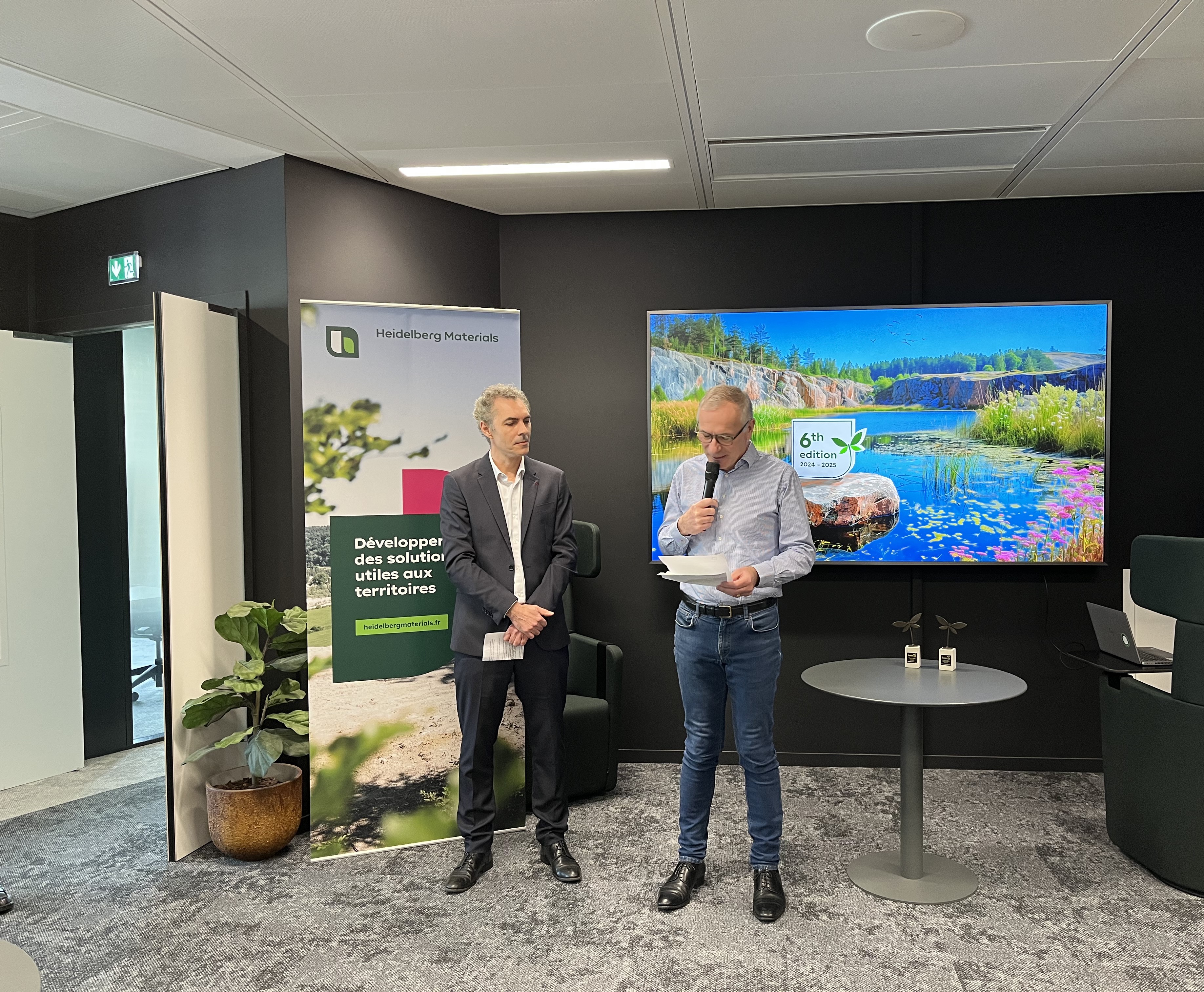 Deux hommes debout dans une salle de conférence moderne. L'homme à gauche porte un costume et se tient à côté d'un panneau avec des illustrations et un logo bancaire. L'homme à droite, en chemise bleue et jeans, tient un micro et semble s'exprimer. En arrière-plan, un écran affiche une image d'un paysage avec un lac, des arbres et des fleurs. Une table basse et des chaises modernes se trouvent à côté.