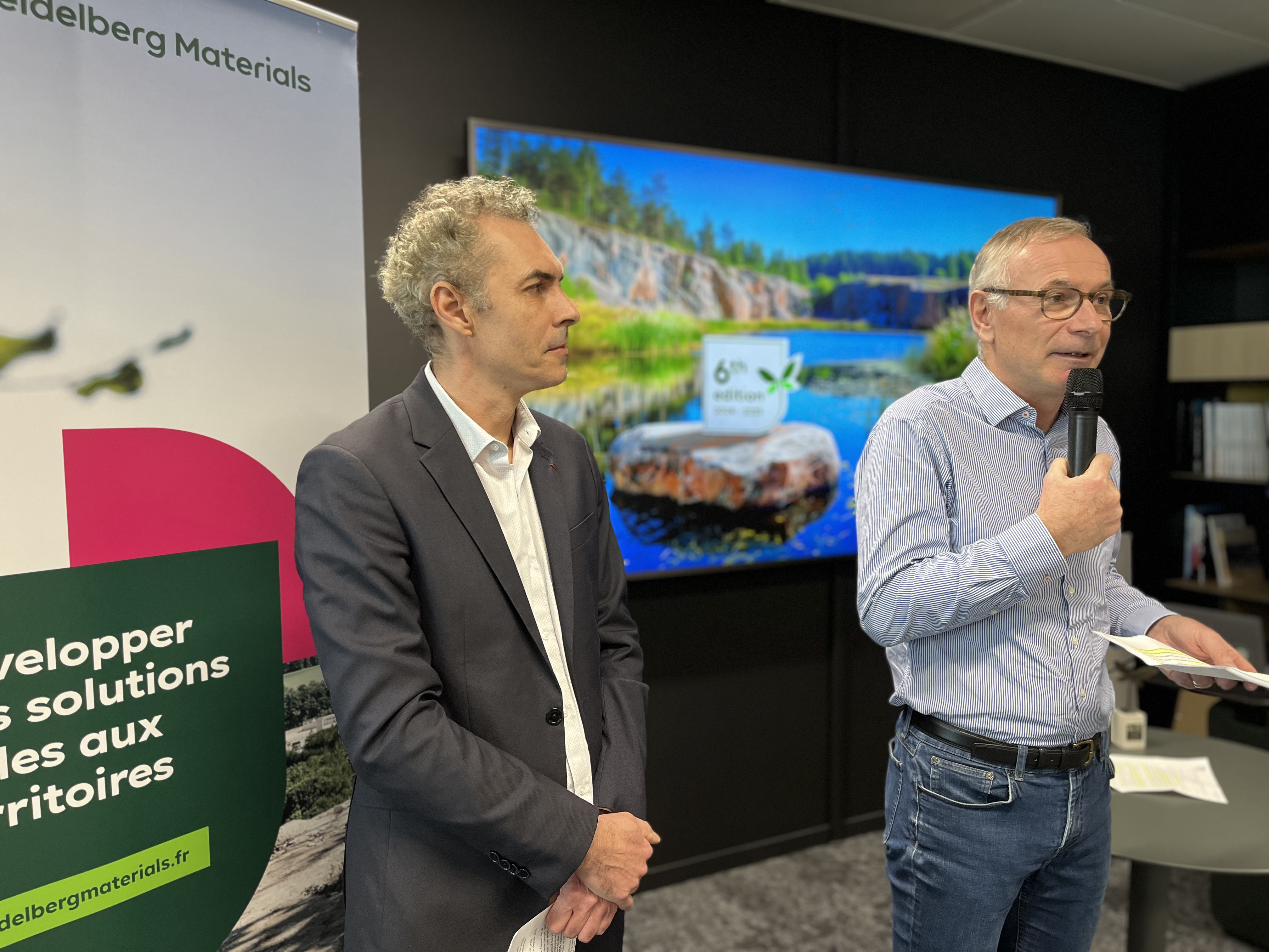 Deux hommes debout dans une salle de conférence. À gauche, un panneau avec le texte « Développer des solutions pour les territoires ». En arrière-plan, un écran affiche un paysage naturel avec un logo au centre. L'homme à droite tient un micro et des feuilles de papier.