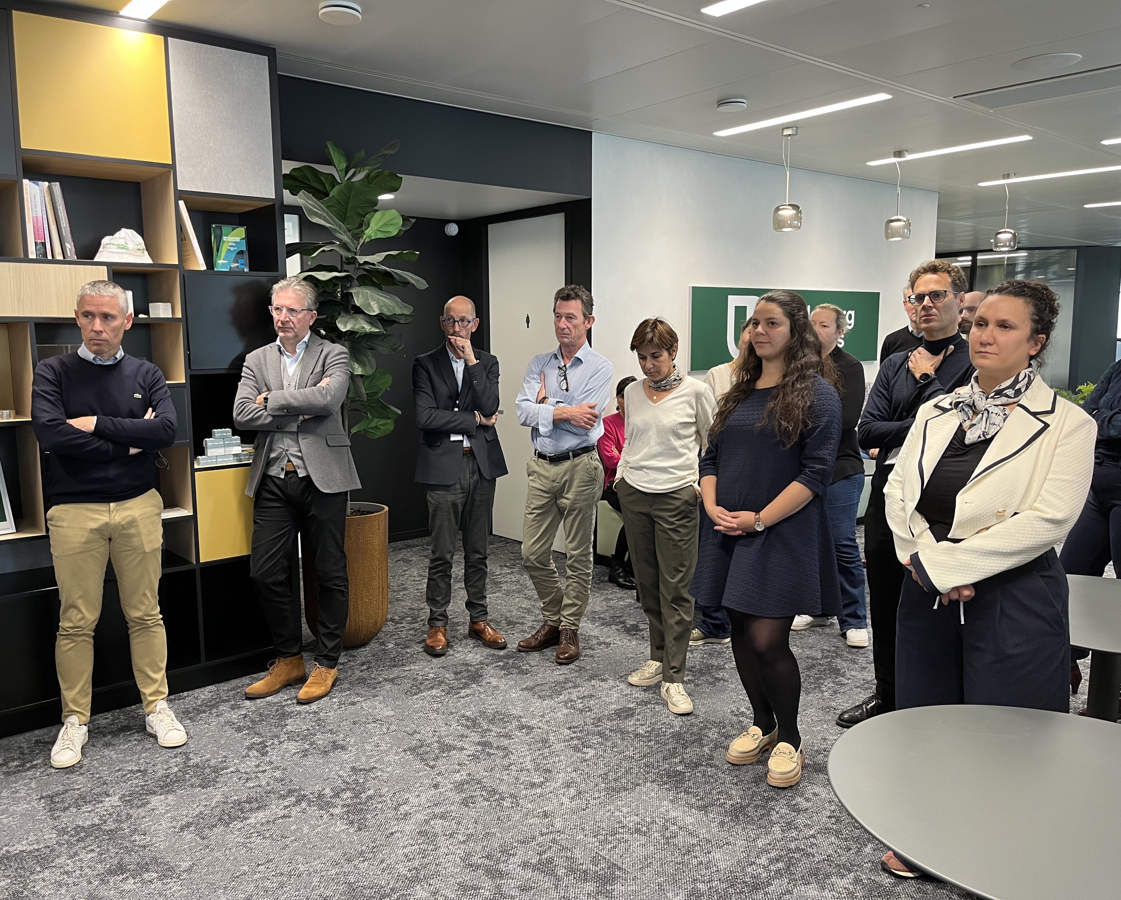 Un groupe de neuf personnes debout dans un espace de bureau moderne, écoutant attentivement une présentation hors champ. À gauche, des étagères décorées avec des livres et des objets, au centre des plantes et des tables rondes. Les participants portent des tenues professionnelles décontractées. L'éclairage est lumineux et l'atmosphère semble studieuse.