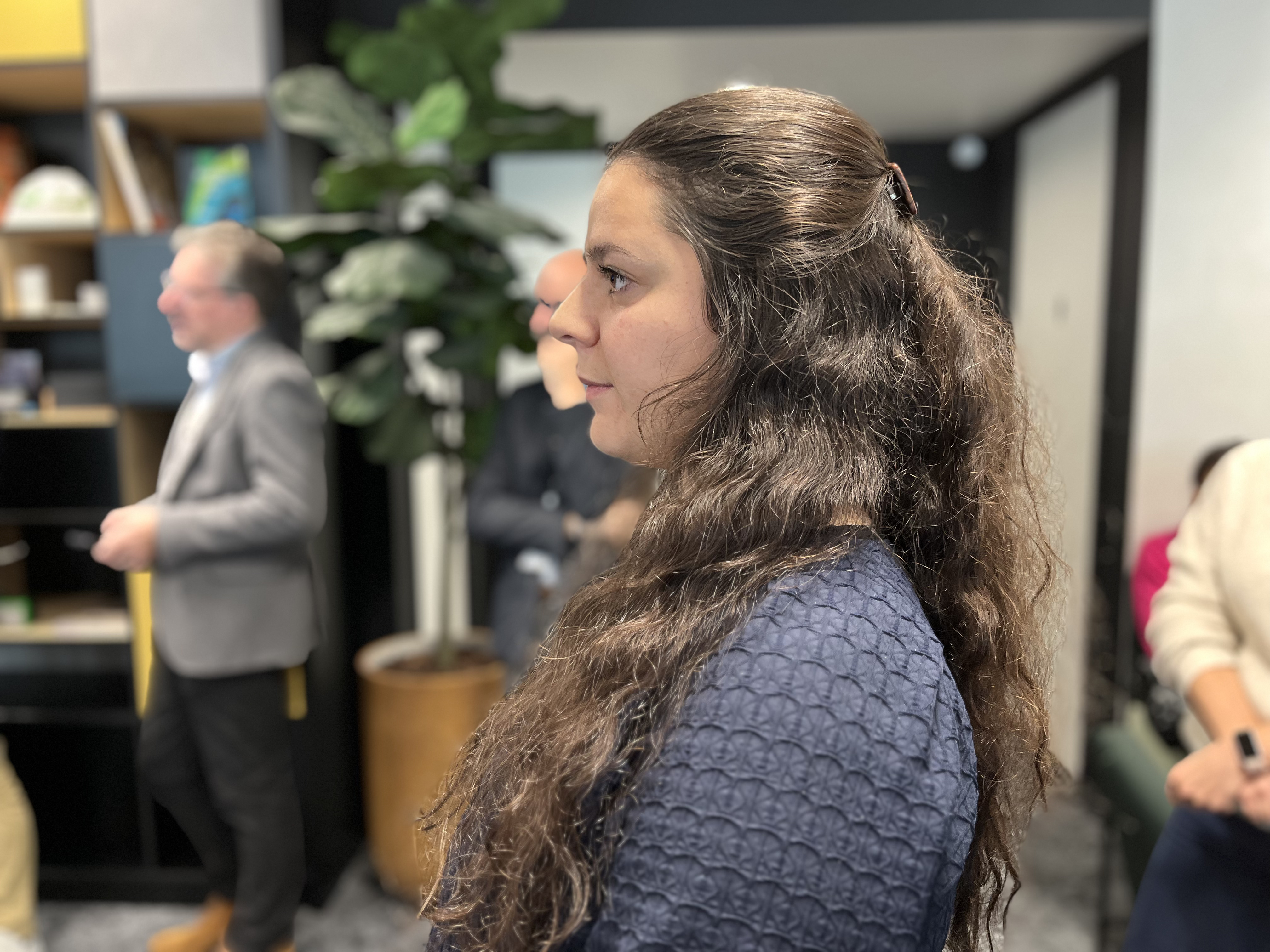 Une personne de dos avec des cheveux longs et bouclés au centre de l'image dans un espace intérieur, entourée de plusieurs autres individus flous en arrière-plan. À gauche, une étagère contenant des livres et des objets décoratifs, et un grand plante verte dans un pot. À droite, une personne avec un pull beige et un mur blanc en arrière-plan.