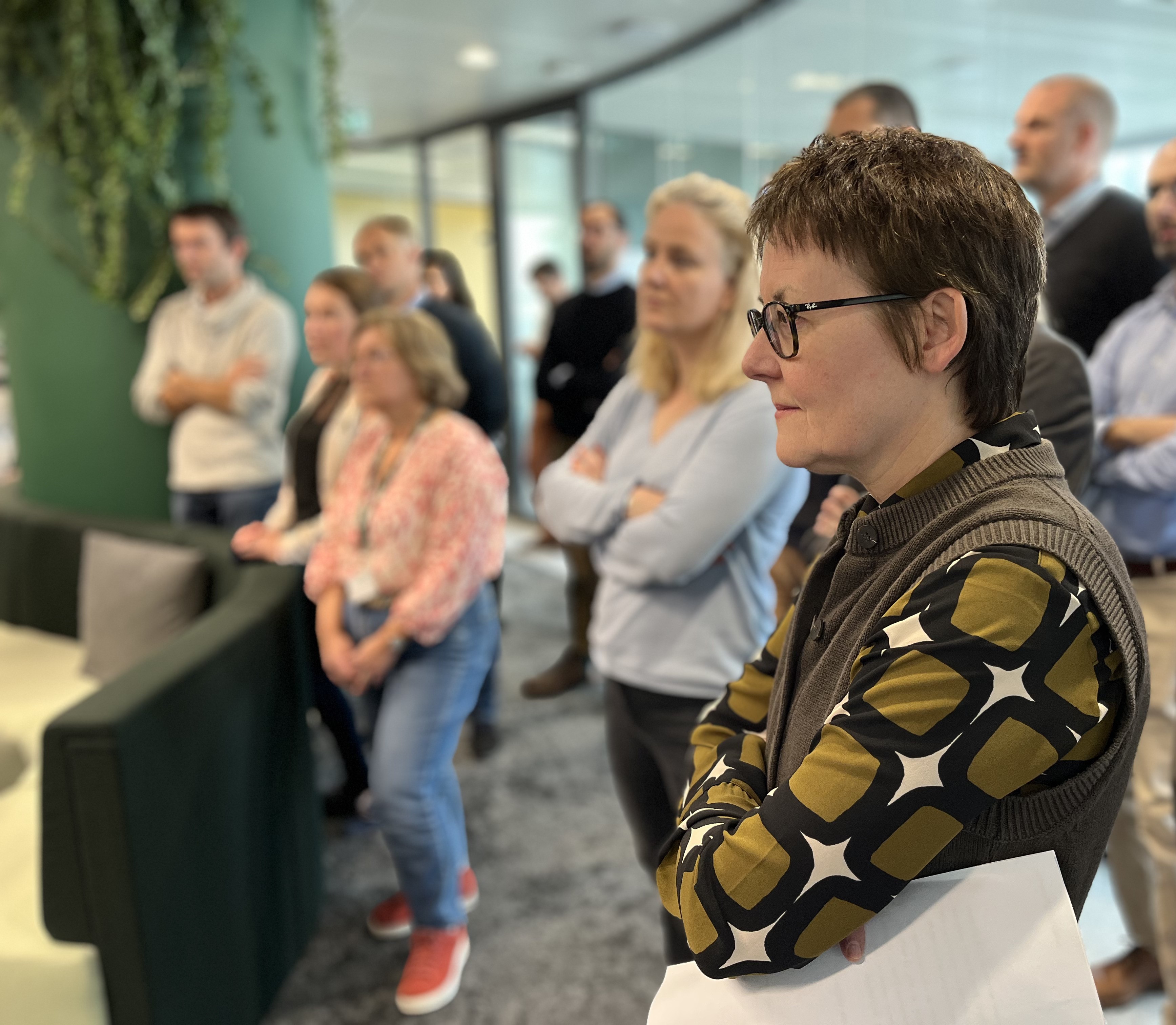 Groupe de personnes debout dans un espace de bureau moderne, écoutant une présentation ou événement. En arrière-plan, des plantes et des éléments de mobilier, avec une ambiance collaborative et attentive.