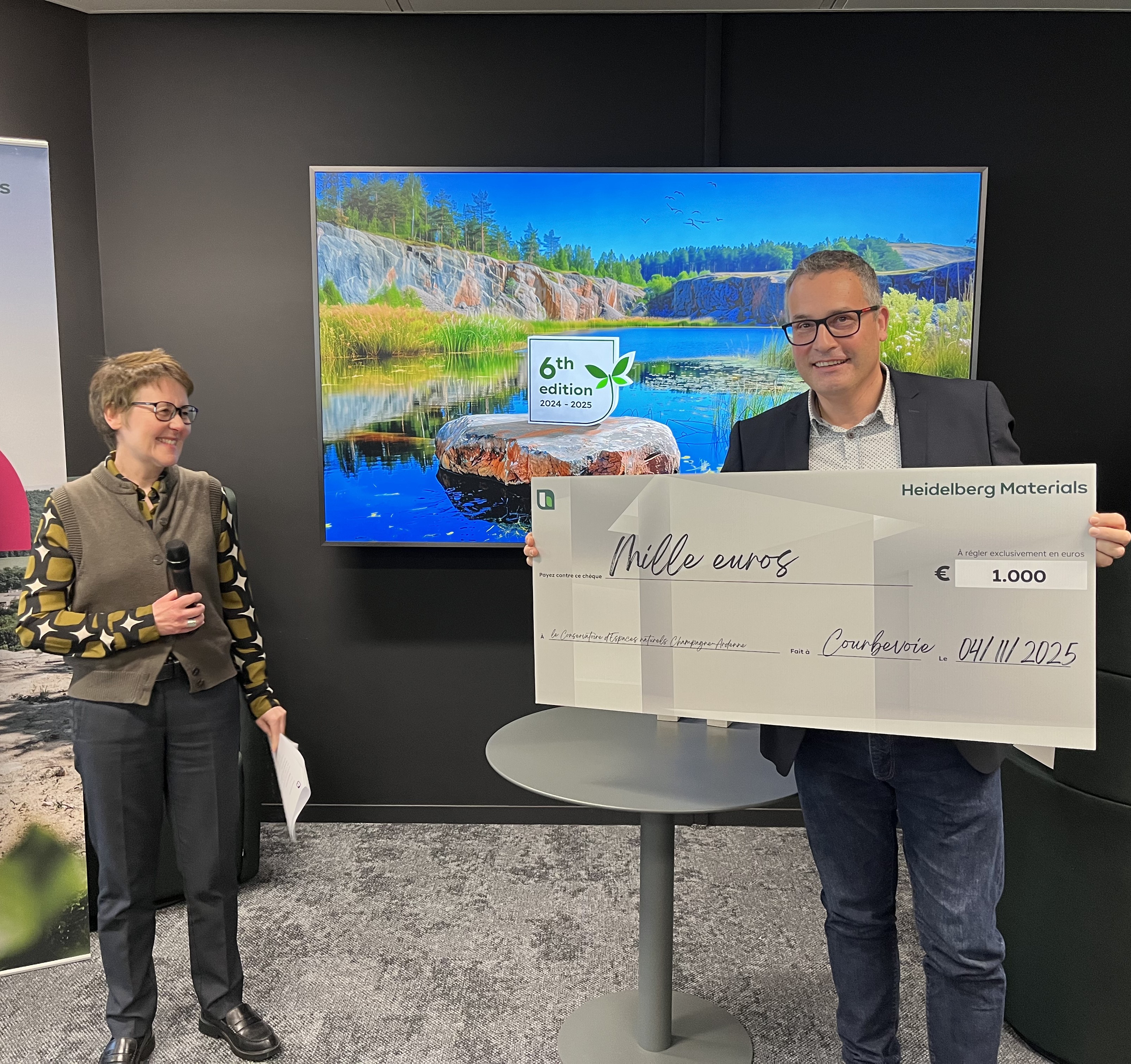 Une femme tenant un microphone se tient à gauche. À droite, une personne tient un grand chèque de 1 000 euros. En arrière-plan, un écran affiche une image naturelle avec un paysage de lac. Une table ronde est visible au centre.