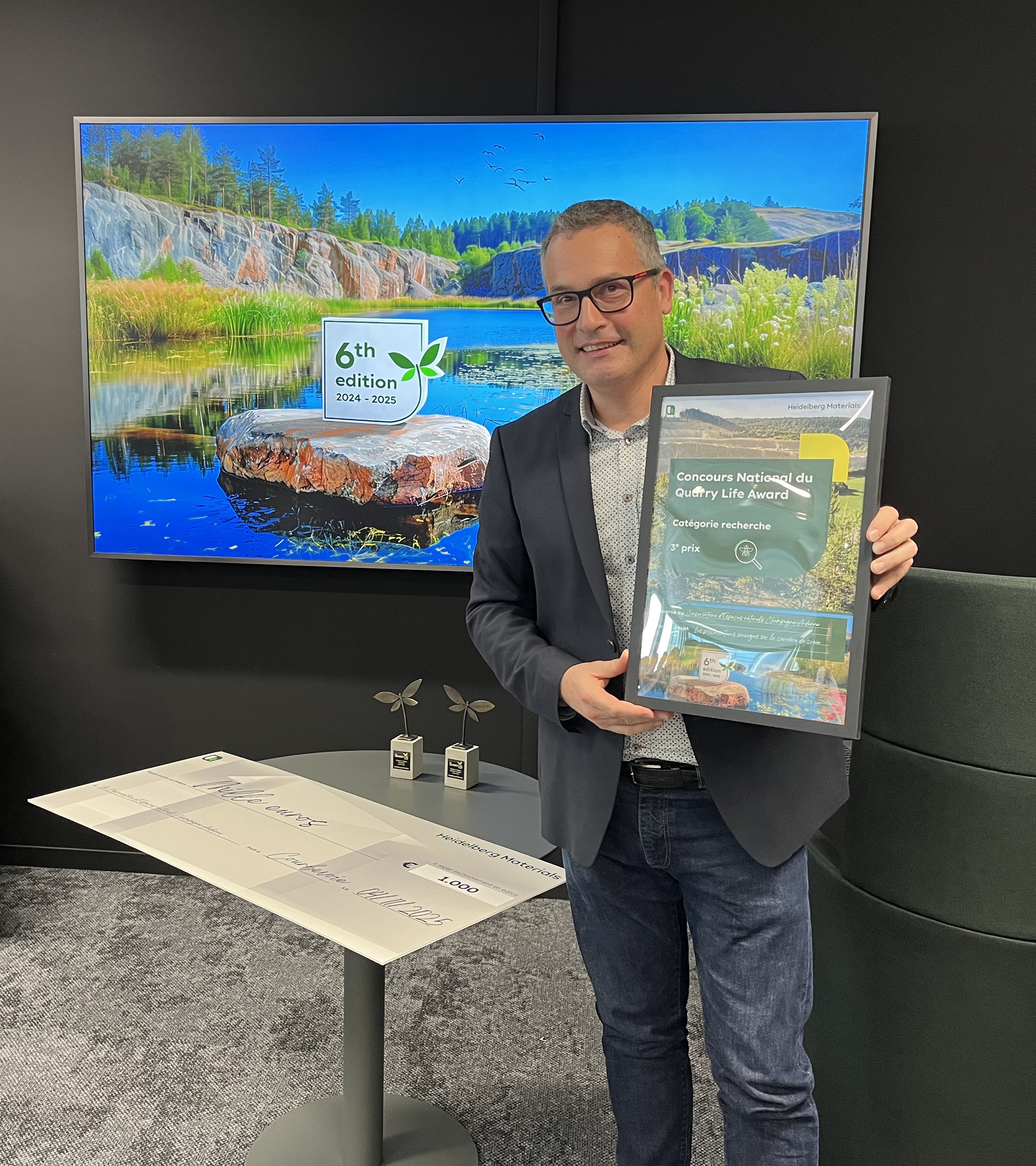 Un homme en costume tient un cadre contenant un document ou un certificat. En arrière-plan, un écran affiche une image d'un paysage naturel avec un rocher, de l'eau, et des arbres, accompagné du texte "6th green week". À gauche, une table avec deux petits trophées posés dessus. L'environnement est professionnel.