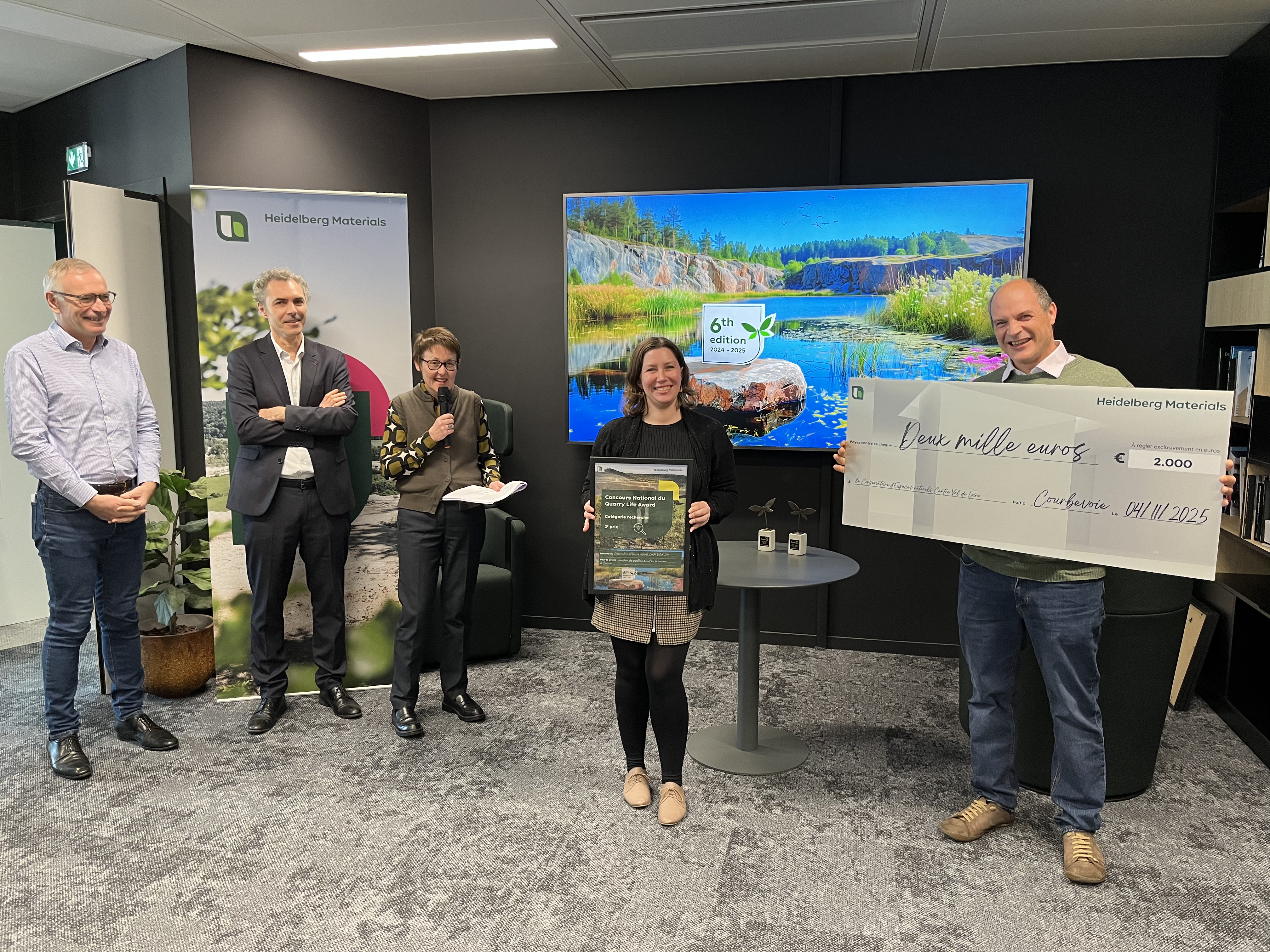 Cinq personnes se tiennent dans une salle de réunion, trois à gauche et deux au centre. Une femme au centre tient un tableau, et un homme à droite tient un grand chèque symbolique. En arrière-plan, un écran affiche une image d'un paysage naturel avec un lac entouré de montagnes.