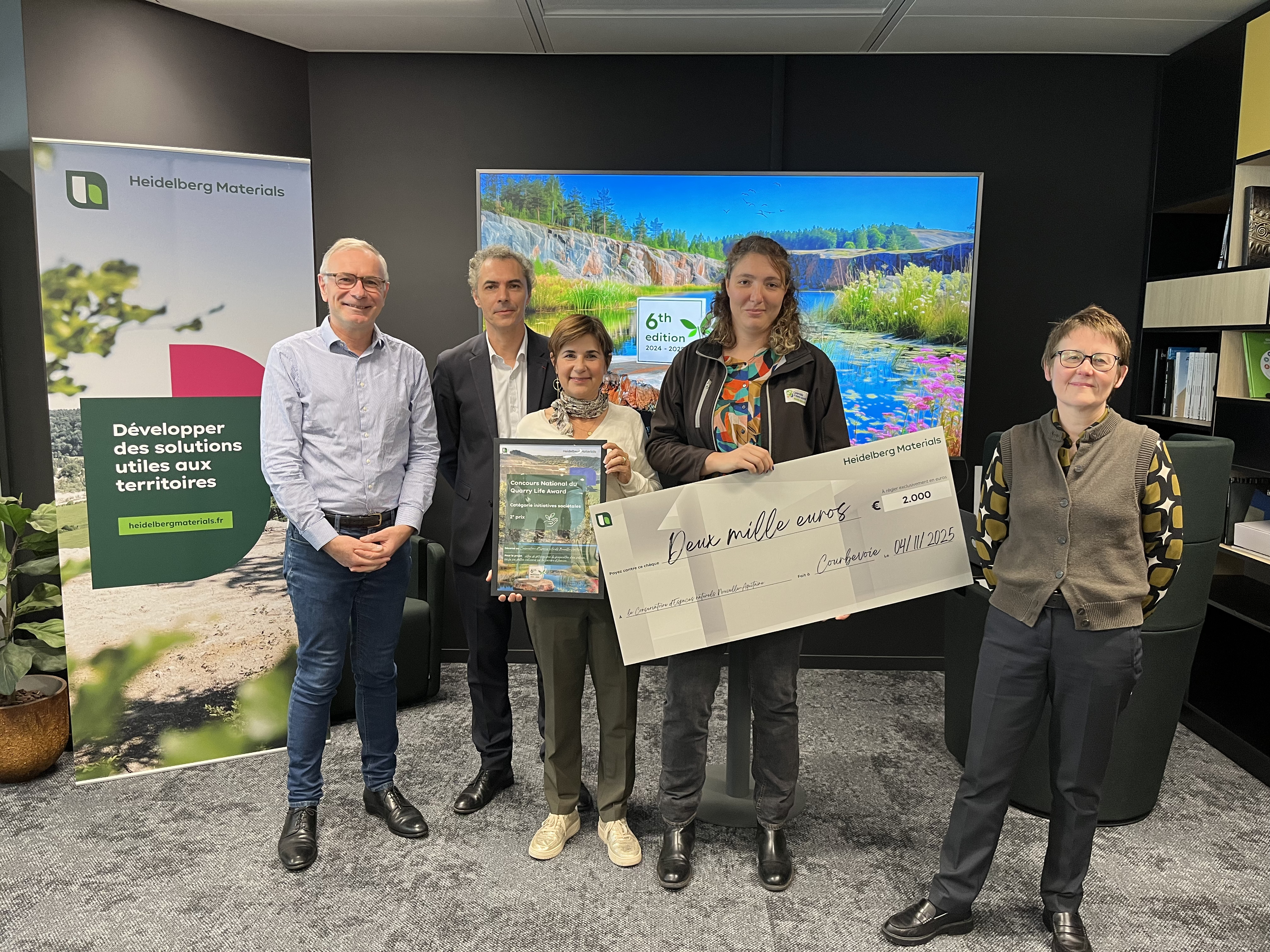 Cinq personnes posent dans un bureau avec un chèque géant de 1000 €. À gauche, un panneau publicitaire d'une organisation. En arrière-plan, un grand écran montre un paysage coloré.