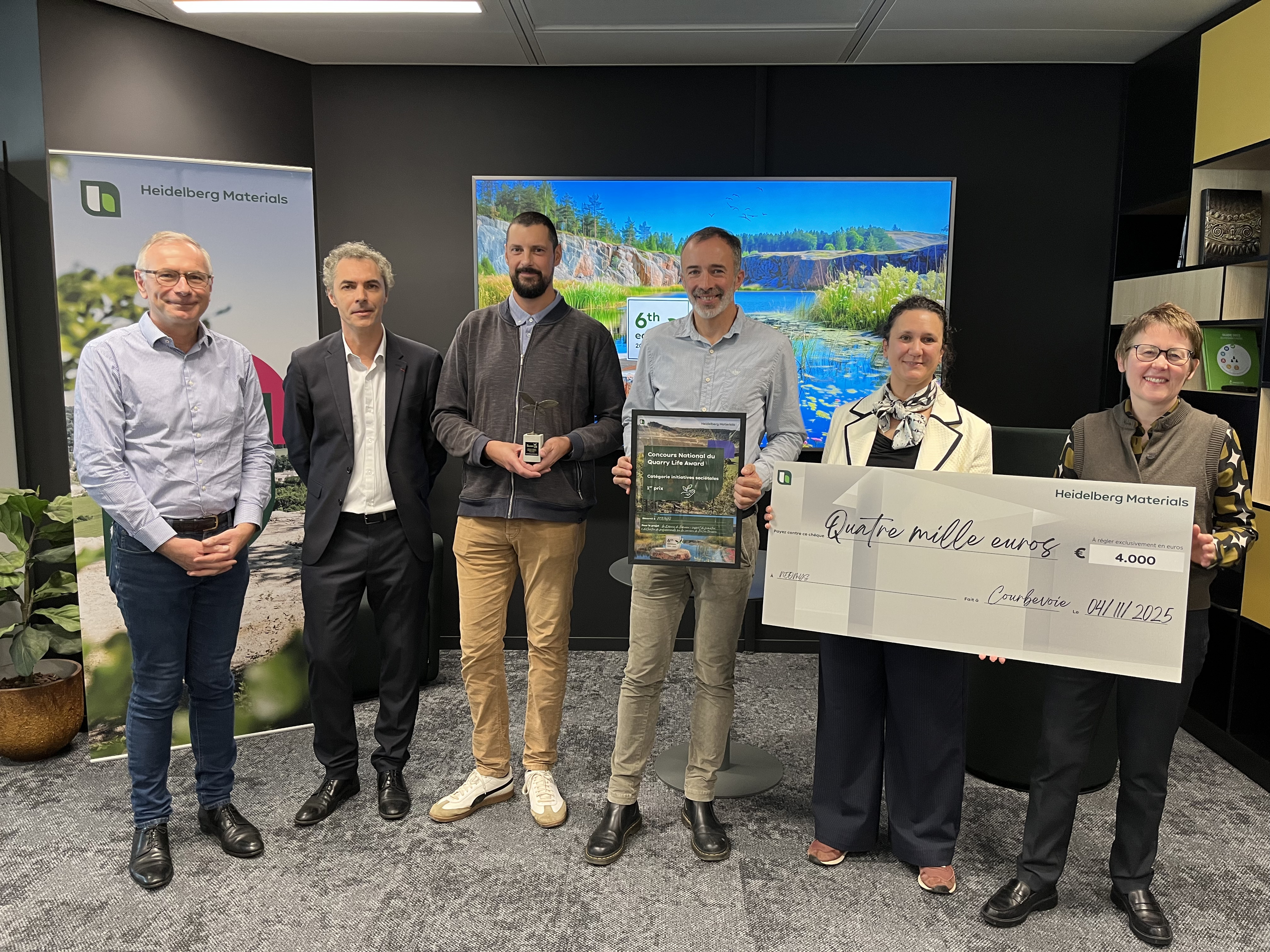 Six personnes posant ensemble dans un environnement professionnel. Trois hommes et trois femmes tiennent un trophée et un chèque symbolique avec la somme de 10 000 € visible. En arrière-plan, une télévision affiche une photographie naturelle avec une rivière et des arbres. Sur la gauche, un panneau indique une organisation environnementale.