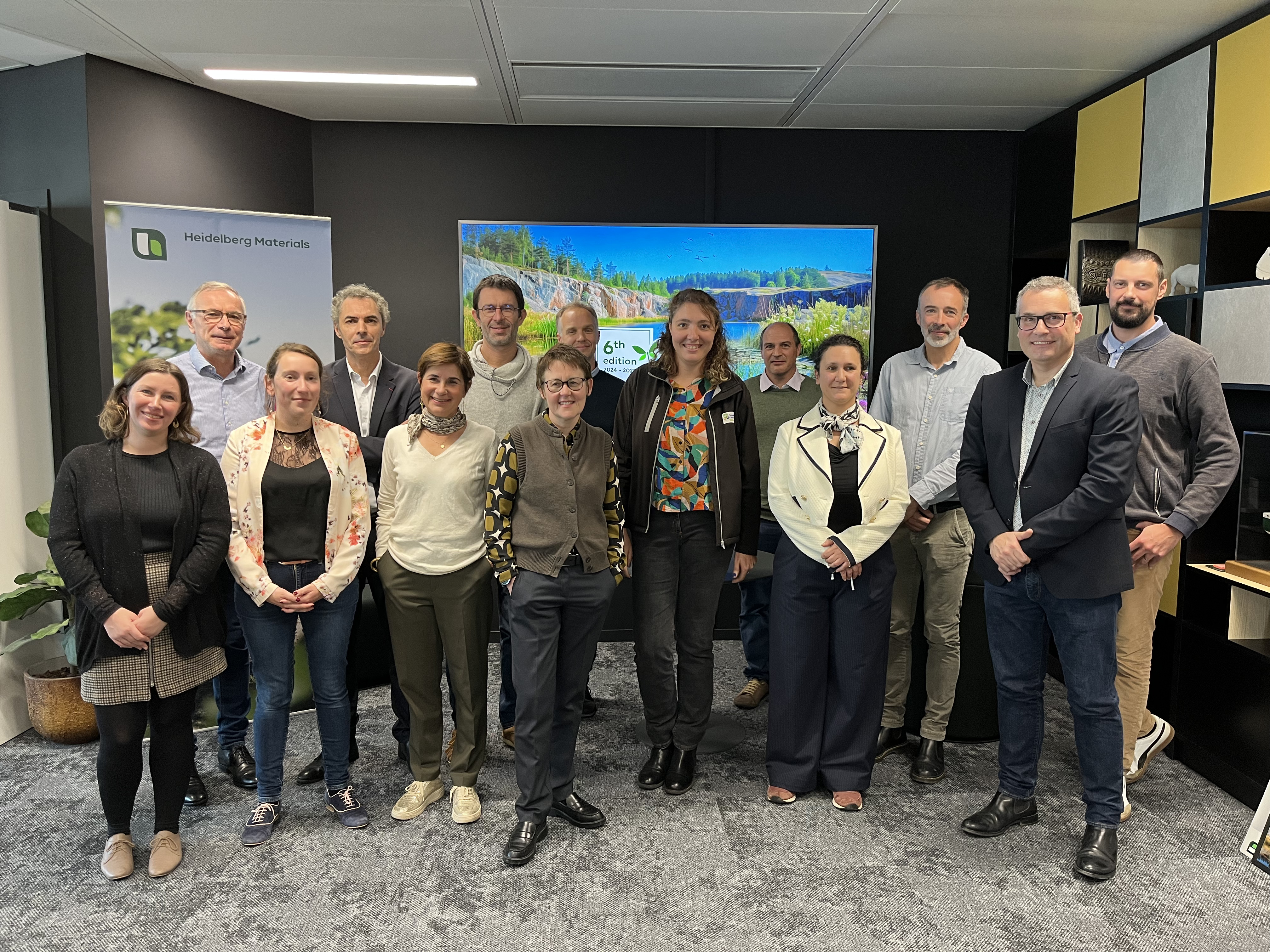 Un groupe de 14 personnes posant ensemble dans un espace de bureau moderne, avec une grande télévision affichant un paysage naturel en arrière-plan, des étagères colorées à droite et une plante à gauche.