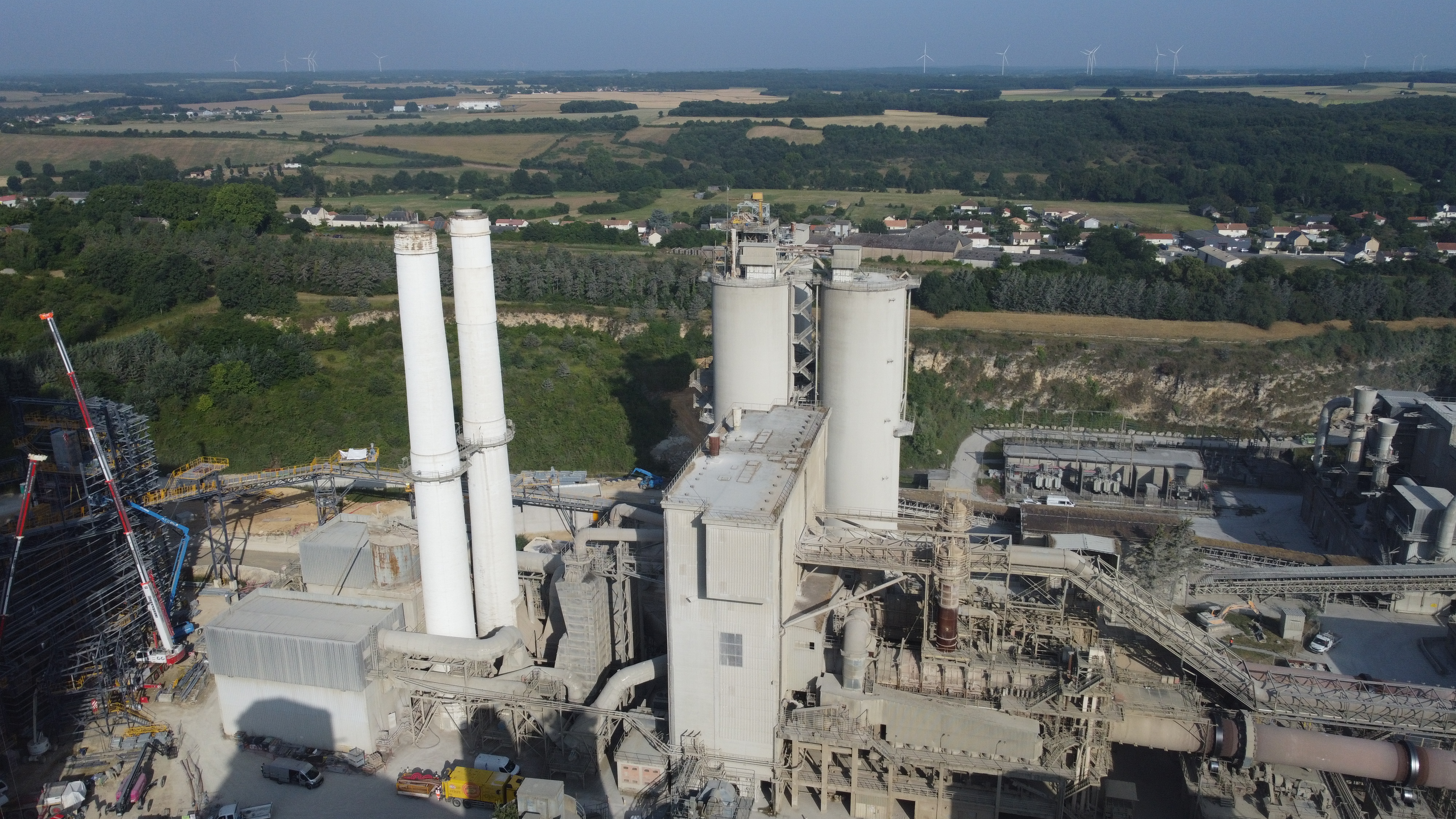 Vue aérienne d'une usine de ciment comprenant deux grandes cheminées blanches au centre, entourées de structures industrielles et de bâtiments annexes. En arrière-plan, on distingue une campagne verdoyante, une zone boisée, et des habitations dispersées.