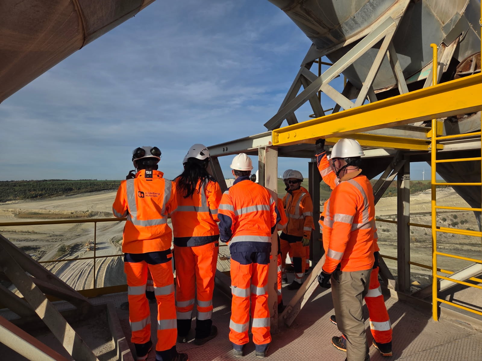 Des ouvriers en tenue de sécurité orange et casques blancs discutent sur un chantier en hauteur, avec une structure métallique et des échafaudages visibles à droite, et un paysage de carrière s'étendant à l'arrière-plan sous un ciel dégagé.