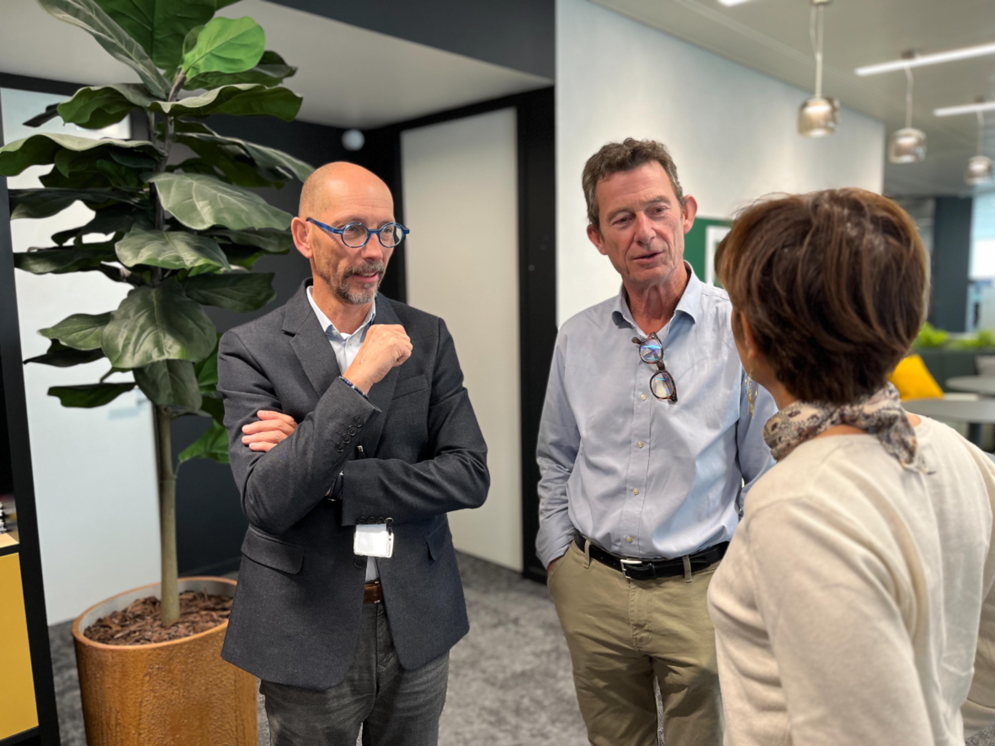 Trois personnes debout discutant dans un espace de bureau moderne avec une plante verte à gauche et une décoration lumineuse en arrière-plan.
