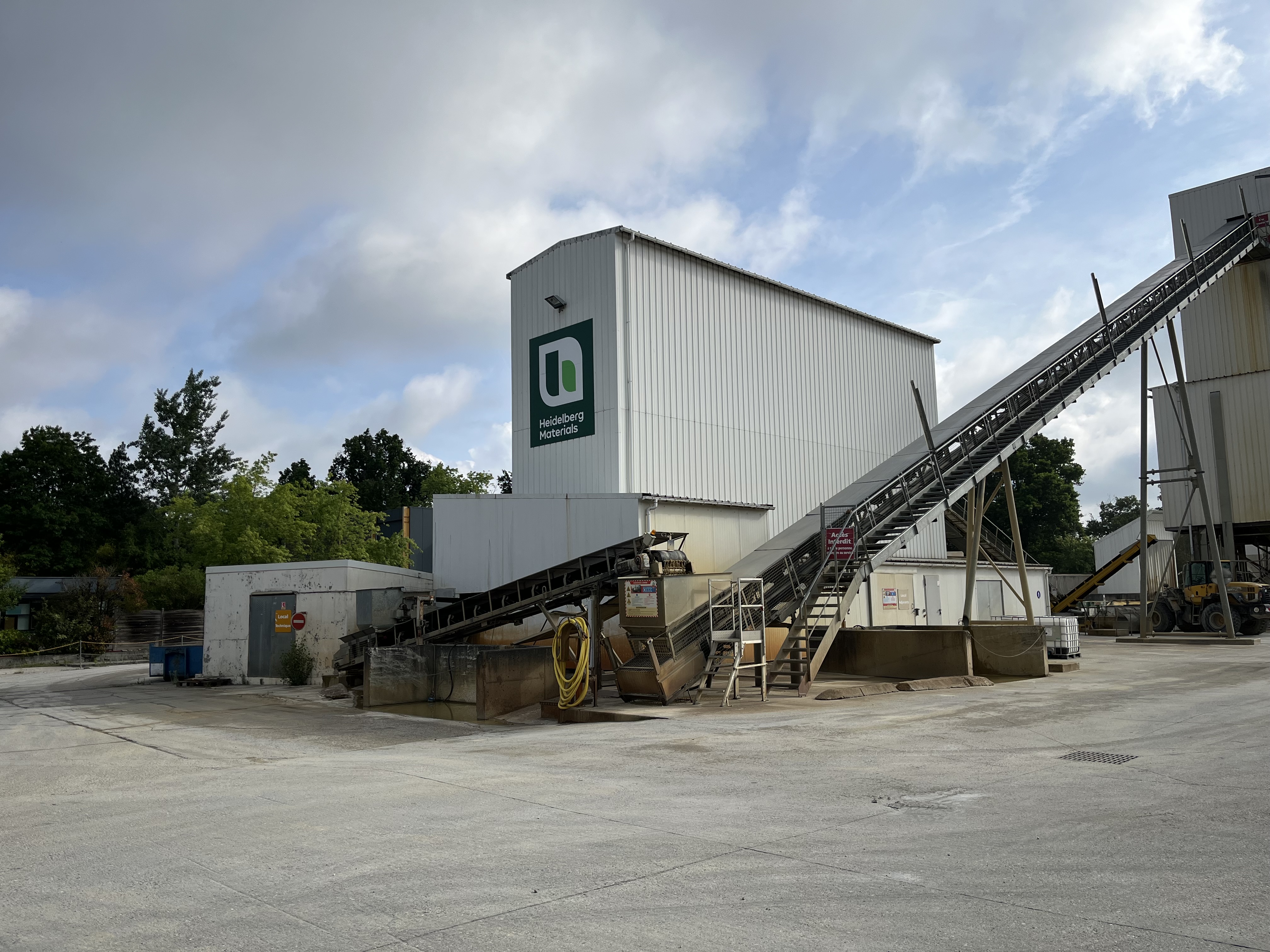 Une centrale à béton avec une tour blanche arborant un logo vert sur sa façade. Des bandes transporteuses s'étendent depuis le côté droit de la structure jusqu'au sol. La centrale est entourée d'arbres et d'un ciel bleu nuageux. Le sol est pavé et exempt de toute activité.