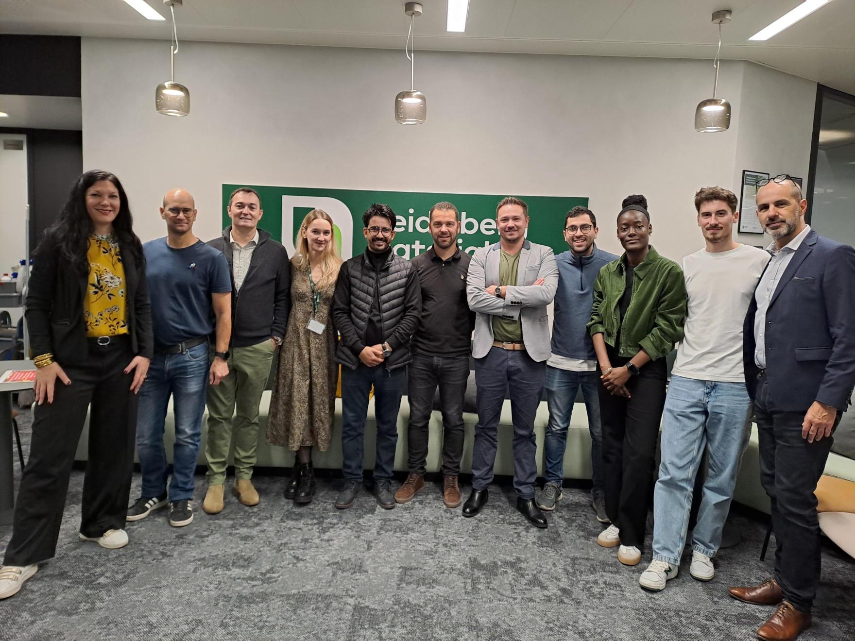 Un groupe de dix personnes posant debout dans une salle de réunion. À l’arrière-plan, un panneau vert avec un texte partiellement lisible ("this is be"). Les individus sont habillés de manière variée, avec un mélange de tenue décontractée et professionnelle. Éclairage doux avec trois suspensions au plafond visibles. Sol en moquette grise.