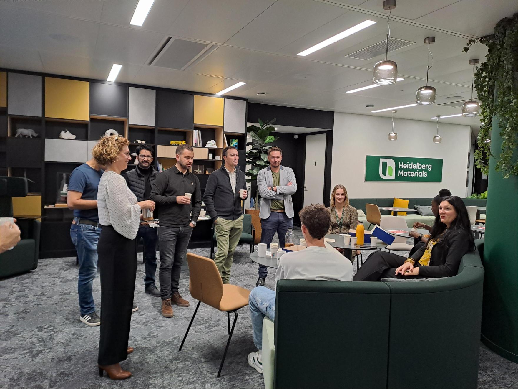 Des employés discutent dans un espace de travail moderne avec des étagères au fond, un panneau "Handelsbanken" vert à droite et des sièges confortables. Une femme debout parle à un groupe debout et assis. L'éclairage est lumineux, et des plantes décorent l'espace.