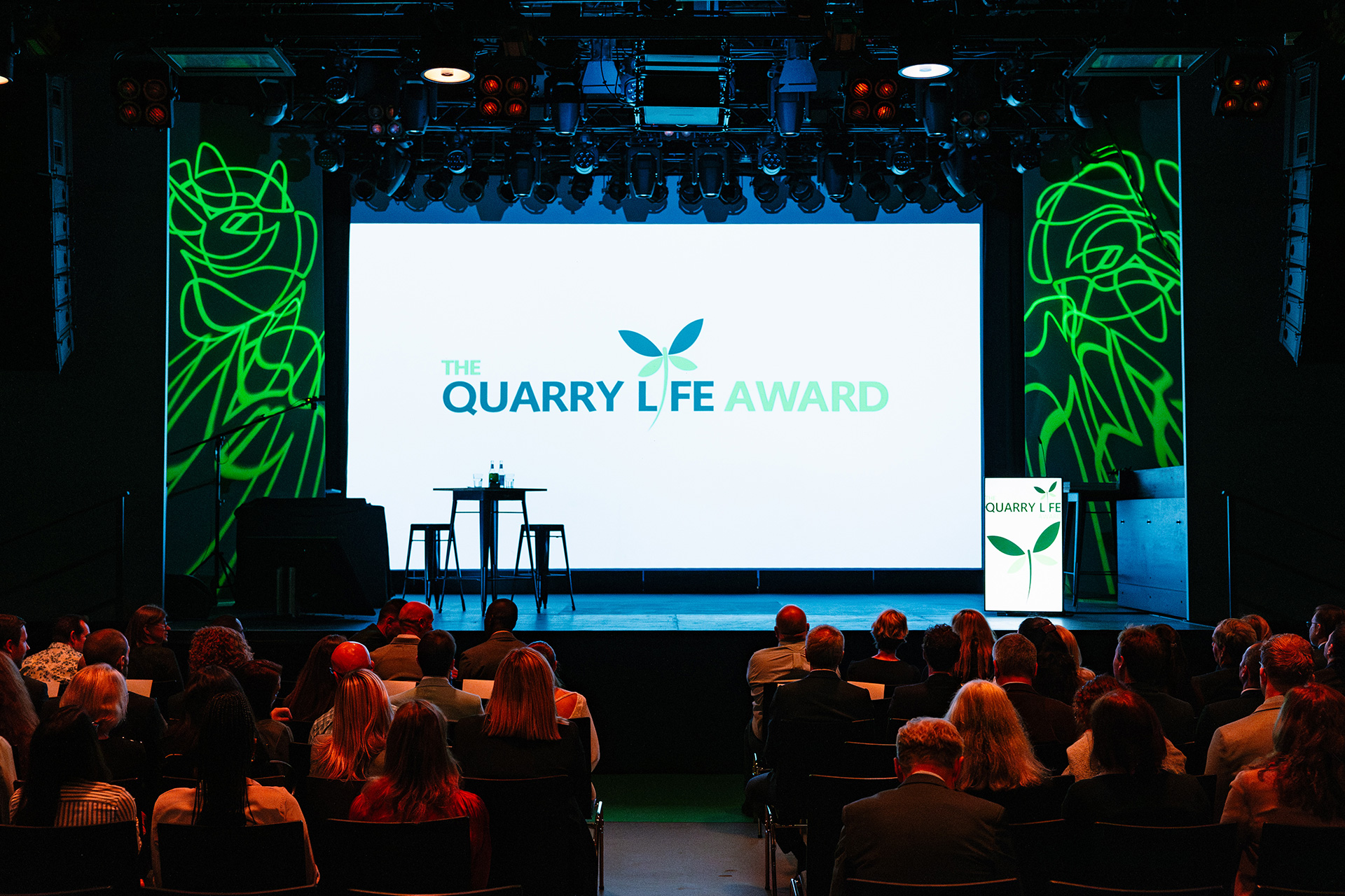 Salle de conférence, vue depuis l’arrière, avec un écran central affichant "The Quarry Life Award" en bleu et vert. Des dessins verts de plantes ornent les murs latéraux. Un pupitre et deux tabourets sont disposés sur scène, à gauche. Des spectateurs, assis en rangées, assistent à l’événement.