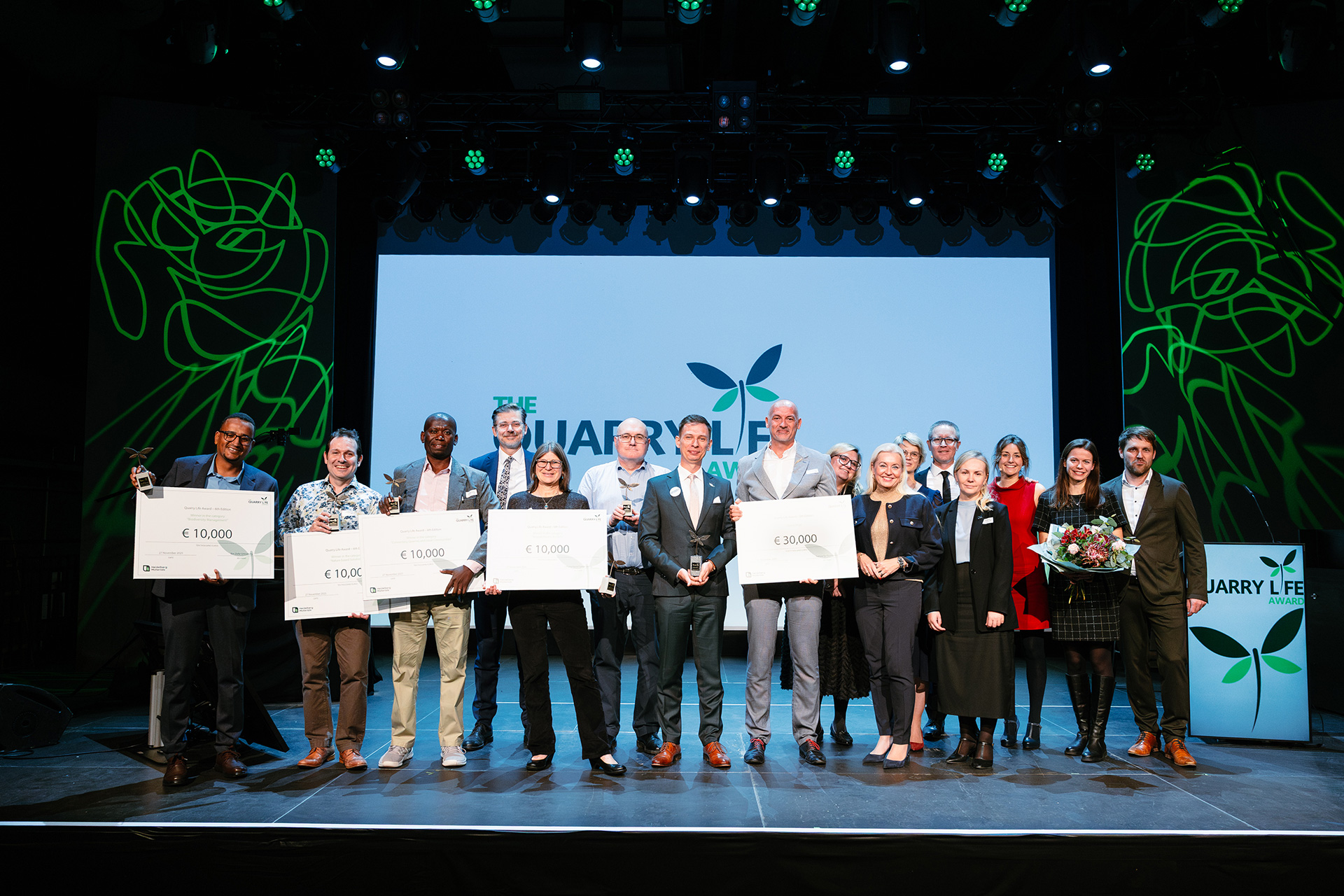 Un groupe de lauréats et organisateurs pose sur scène avec des chèques et un trophée lors d’une remise de prix. En arrière-plan, un grand écran affiche le logo et le nom de l’événement avec des décorations vertes sur les côtés. Les participants sourient, vêtus de tenues professionnelles.