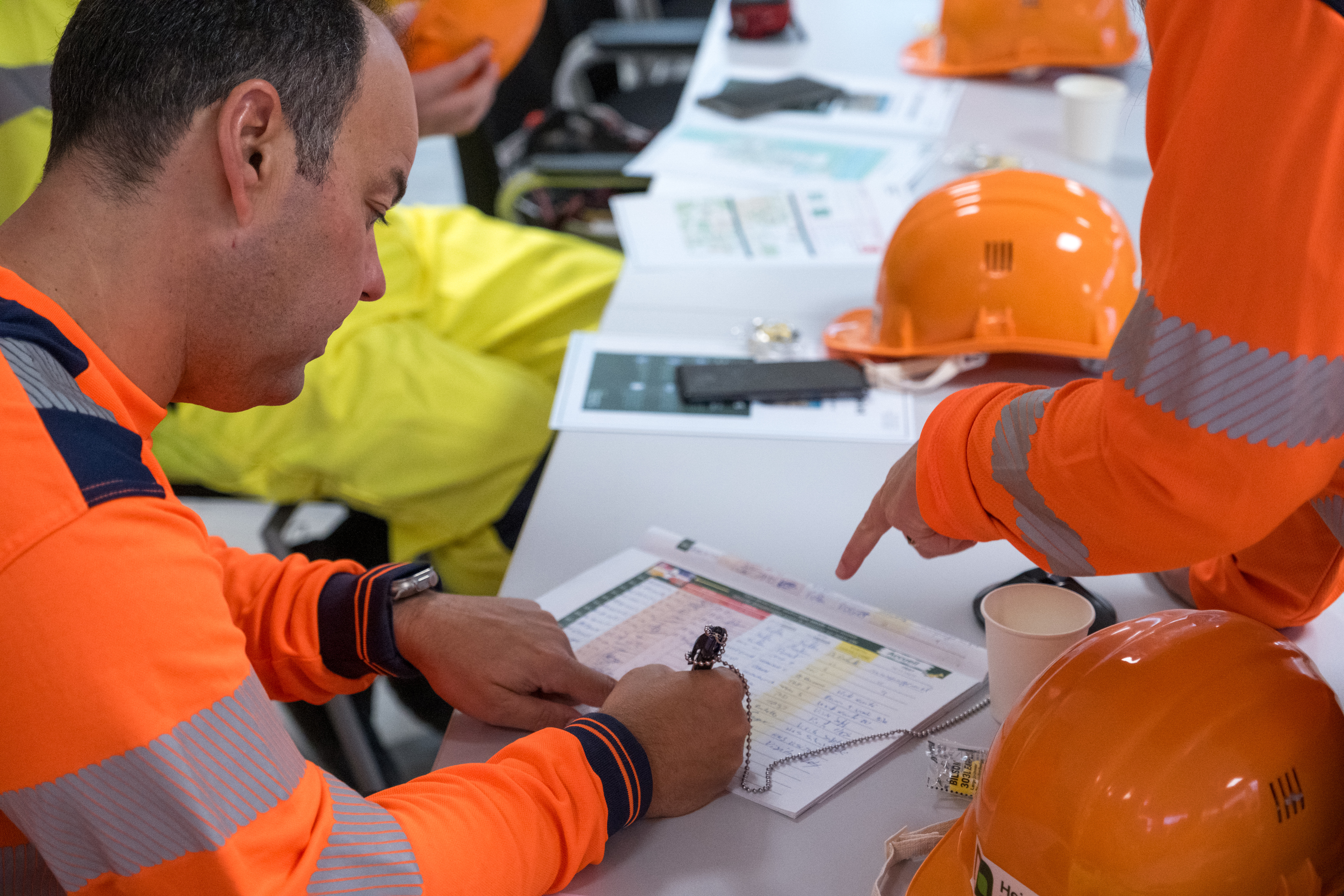Un ouvrier en tenue de sécurité orange remplit un formulaire à une table, tandis qu'une autre personne en uniformes similaires pointe un document. Plusieurs casques de sécurité orange, des documents et un gobelet en carton se trouvent sur la table.