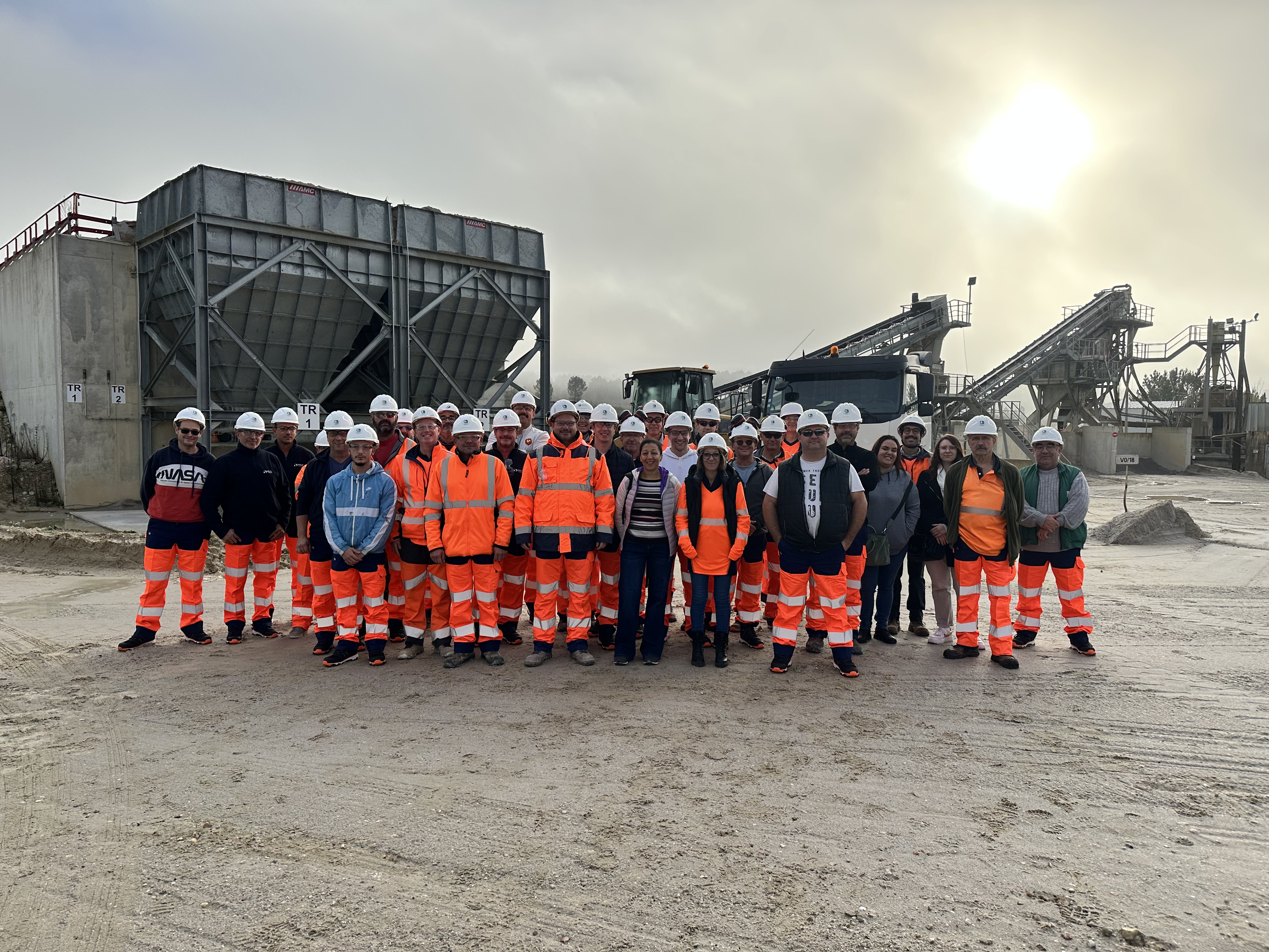 Groupe de travailleurs portant des équipements de sécurité, notamment des casques et des vêtements haute visibilité, posant devant une installation industrielle avec des silos et des machines de traitement des matériaux sous un ciel partiellement ensoleillé.