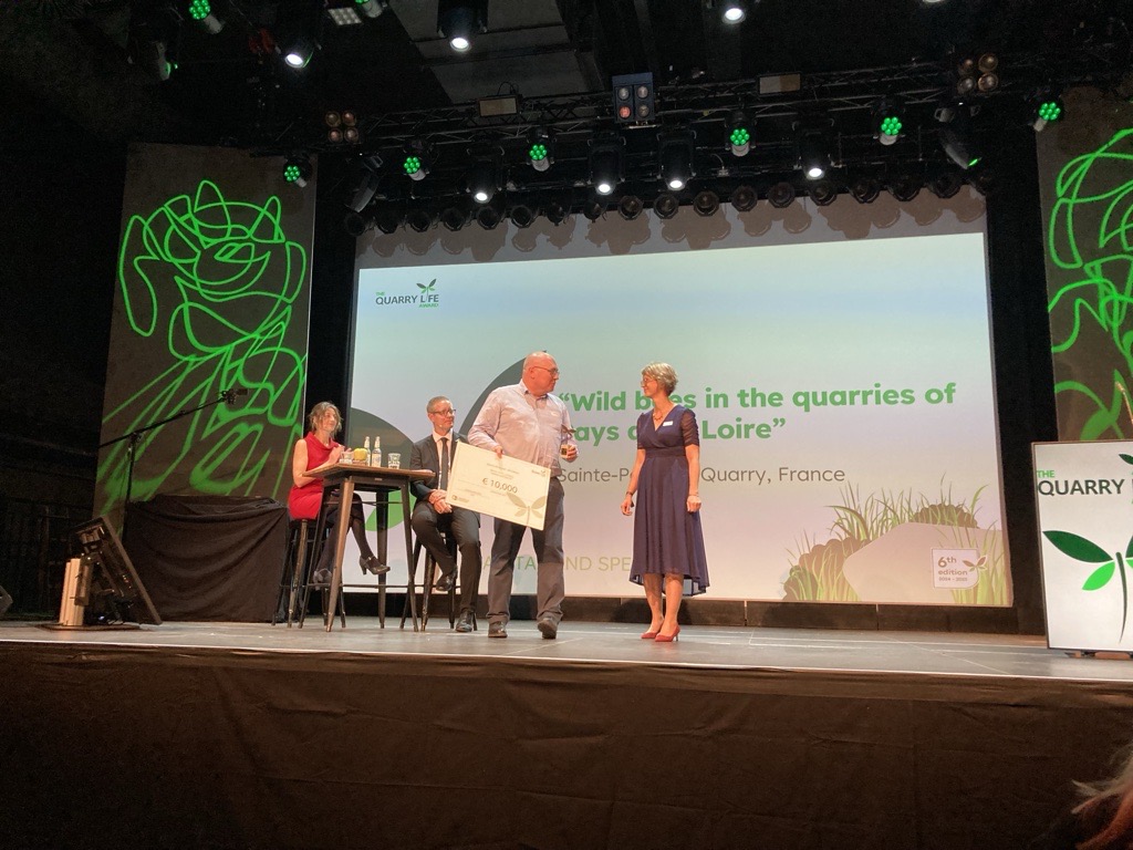 Cérémonie de remise de prix sur scène avec quatre intervenants. Une femme en robe bleue et un homme en chemise blanche tiennent un certificat. En arrière-plan, une présentation projette le titre "Biodiversity in the quarries of Pays de la Loire", accompagné de logos. Décor sur le thème de la nature avec des motifs verts.