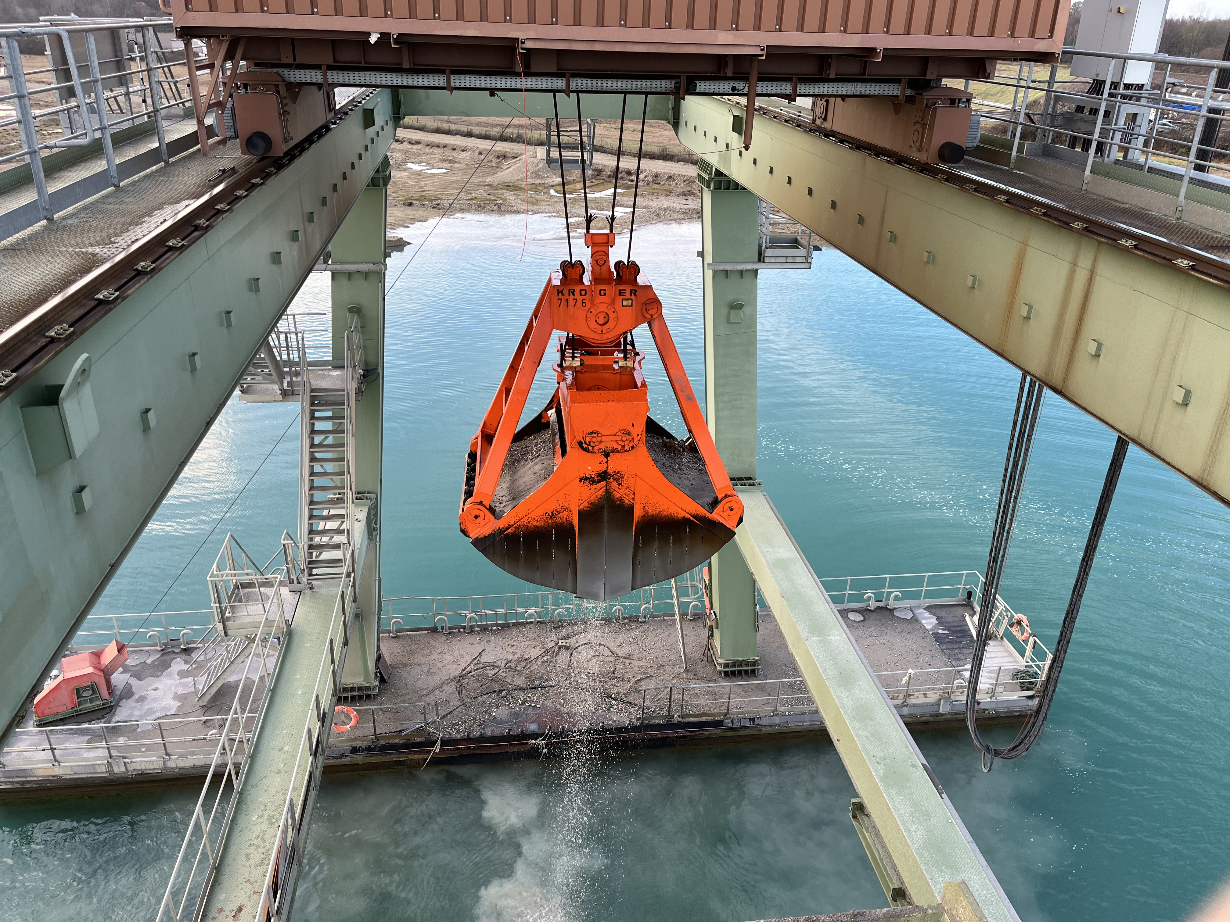 Une benne industrielle orange suspendue à une grue déverse son contenu dans une péniche amarrée sur un plan d'eau. La scène est encadrée par des structures et des poutres en acier, avec l'eau visible en arrière-plan.