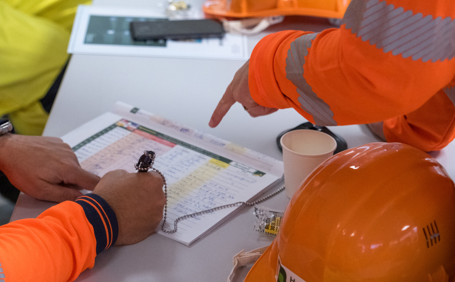 Des travailleurs en gilets de sécurité orange examinent et pointent un formulaire posé sur une table. Un casque de sécurité orange, un stylo et une tasse en papier sont visibles à proximité.