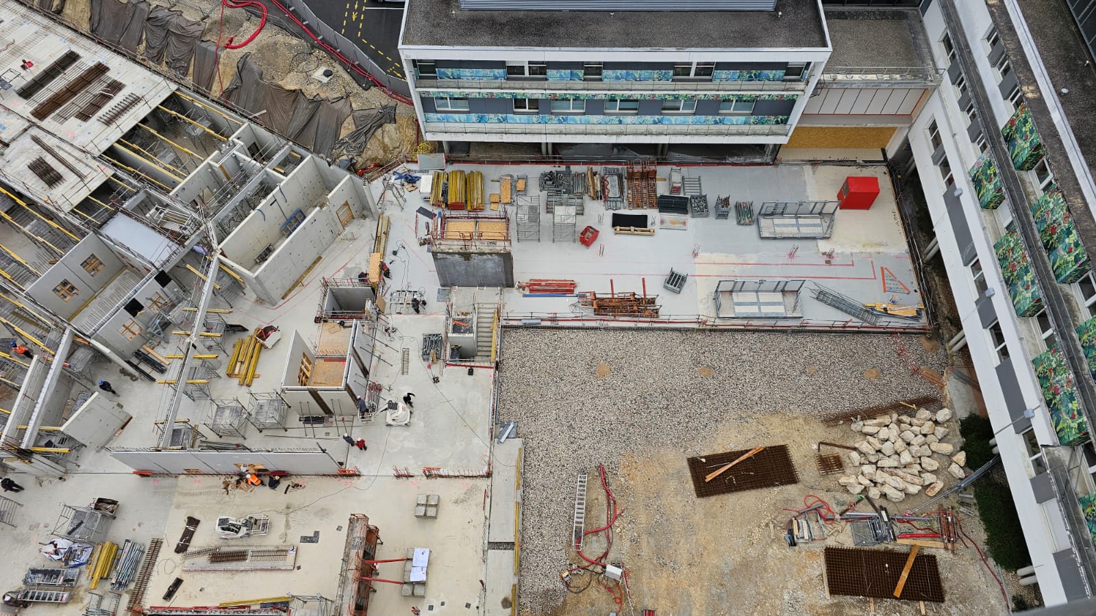Vue aérienne d'un chantier de construction, montrant des structures bétonnées incomplètes à gauche, une zone centrale dégagée avec du gravier et des matériaux de construction, une excavation au bas à droite, et un bâtiment moderne achevé en haut. Des équipements de chantier et des zones de travail balisées sont visibles.