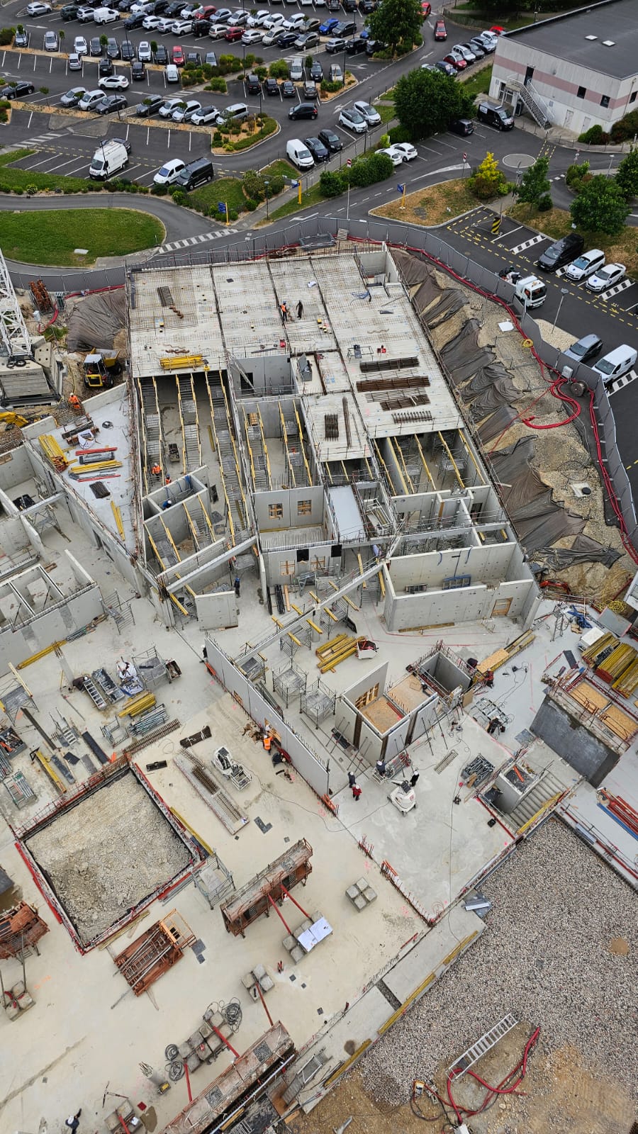 Chantier de construction vu d’en haut, montrant des fondations et structures en béton en cours d’assemblage. Des ouvriers sont présents à différents endroits du site. En arrière-plan, un parking et des espaces verts sont visibles.