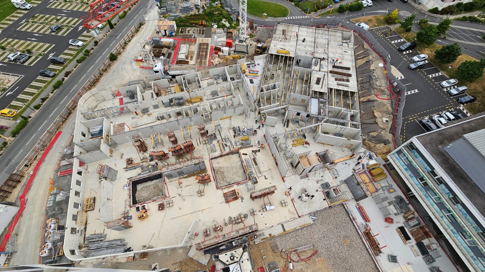 Vue aérienne d'un chantier de construction vaste et en cours, montrant des structures en béton, des poutres, et divers matériaux de construction dispersés. Plusieurs zones désignent des fondations en cours, avec des échafaudages et équipements sur le site. Routes, parkings, et espaces verts visibles autour du chantier.